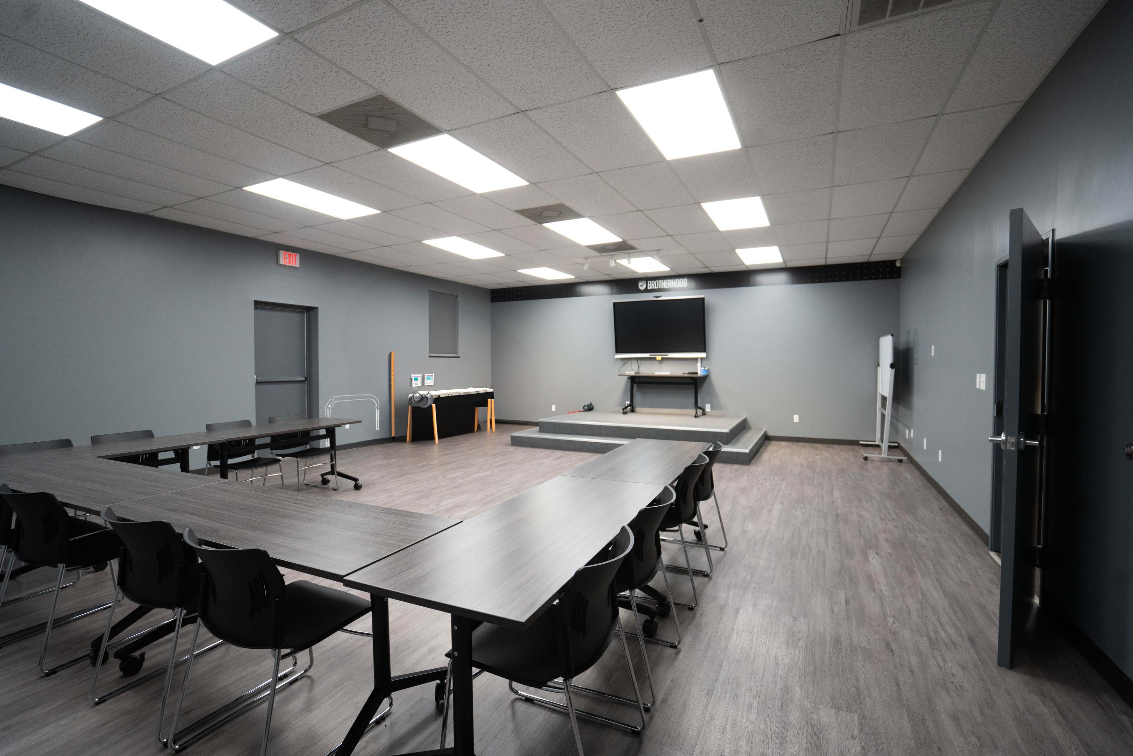 The image shows a spacious, modern meeting room with gray walls, multiple tables arranged in a U-shape, and a stage area at the front.