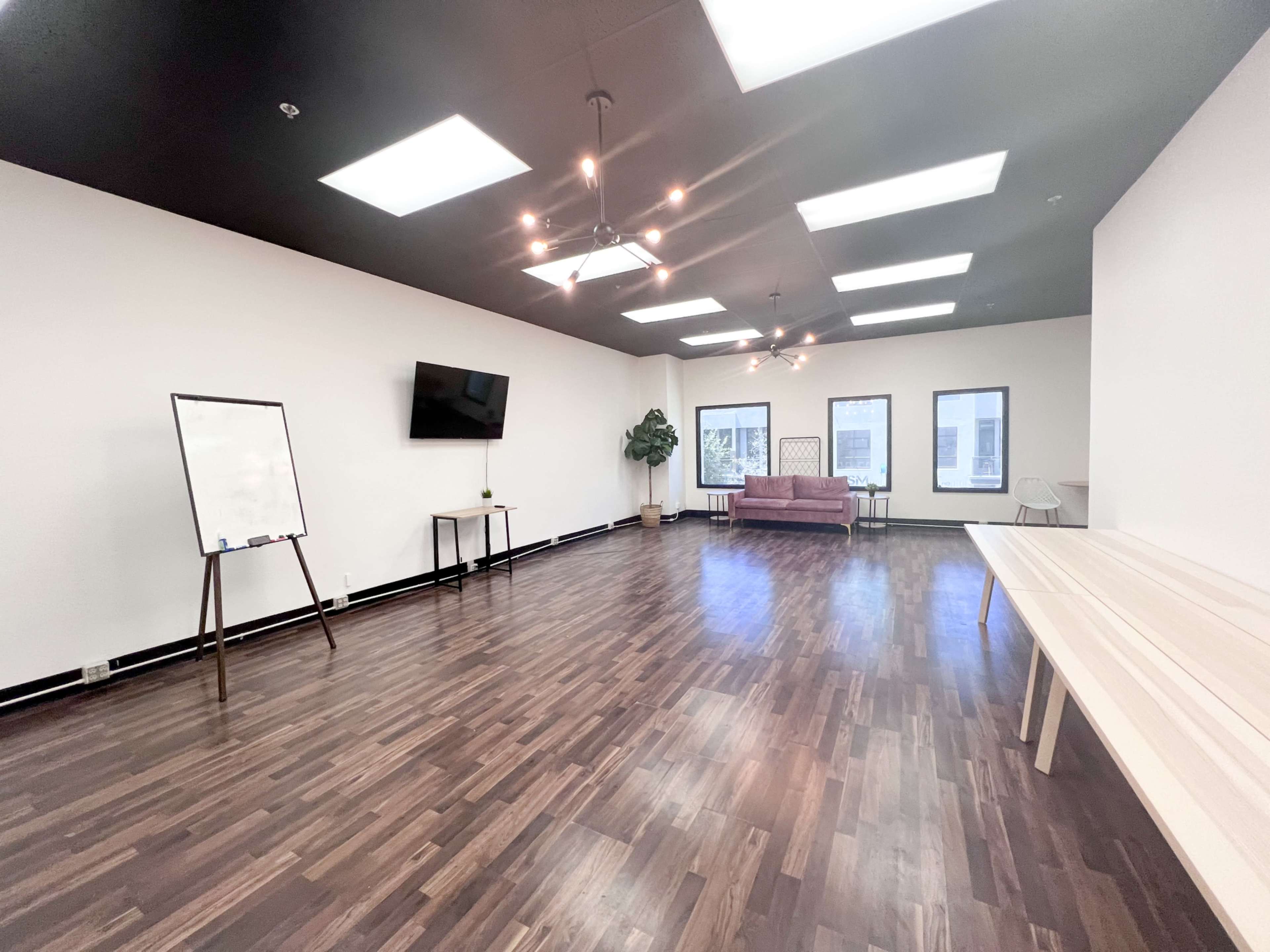 A spacious interior room features a large wooden floor, a television on the wall, a whiteboard stand, a couch, and several windows.