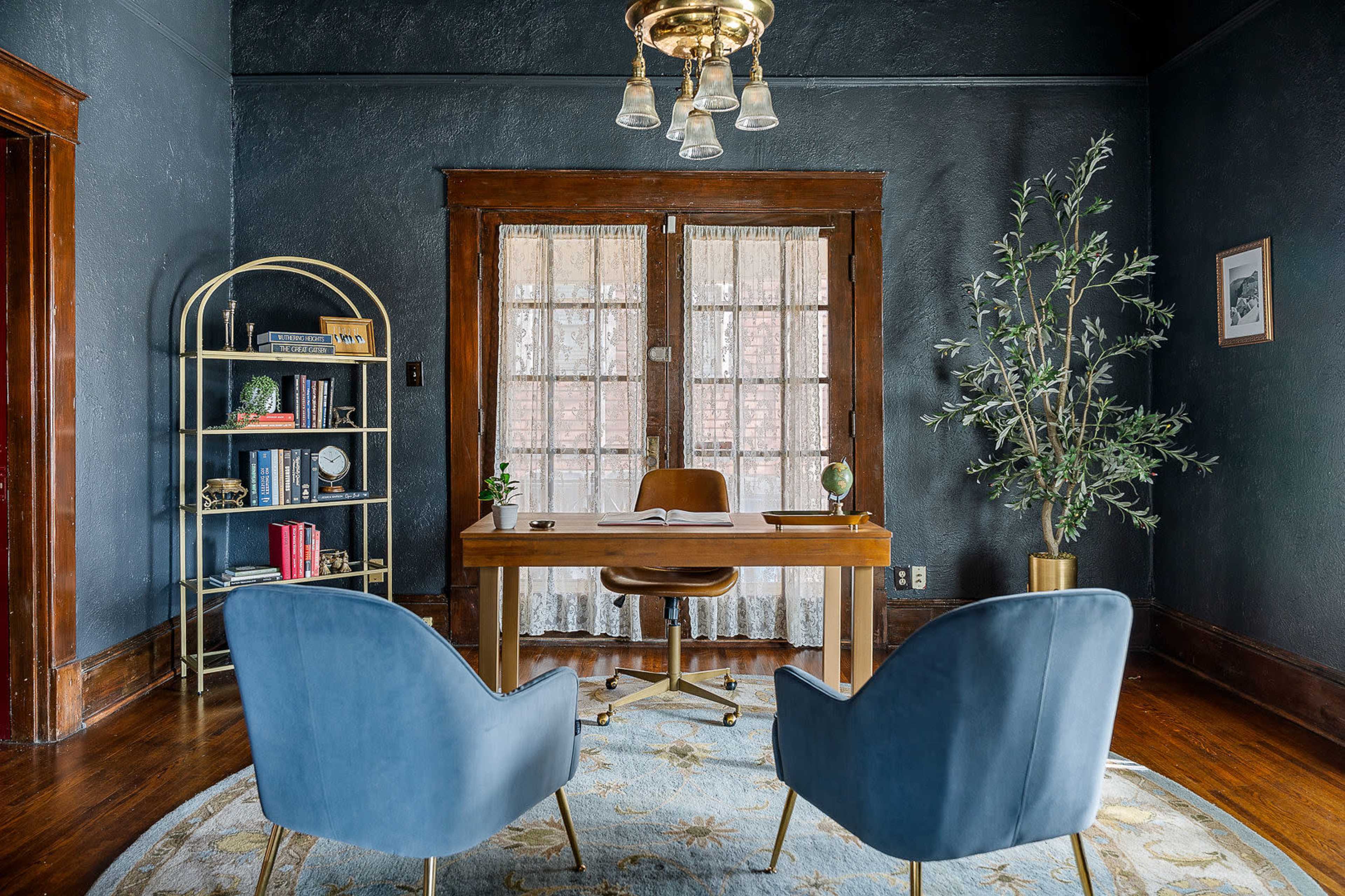 The image shows a home office with a wooden desk, two blue chairs, a gold bookshelf, and large windows covered by sheer curtains.