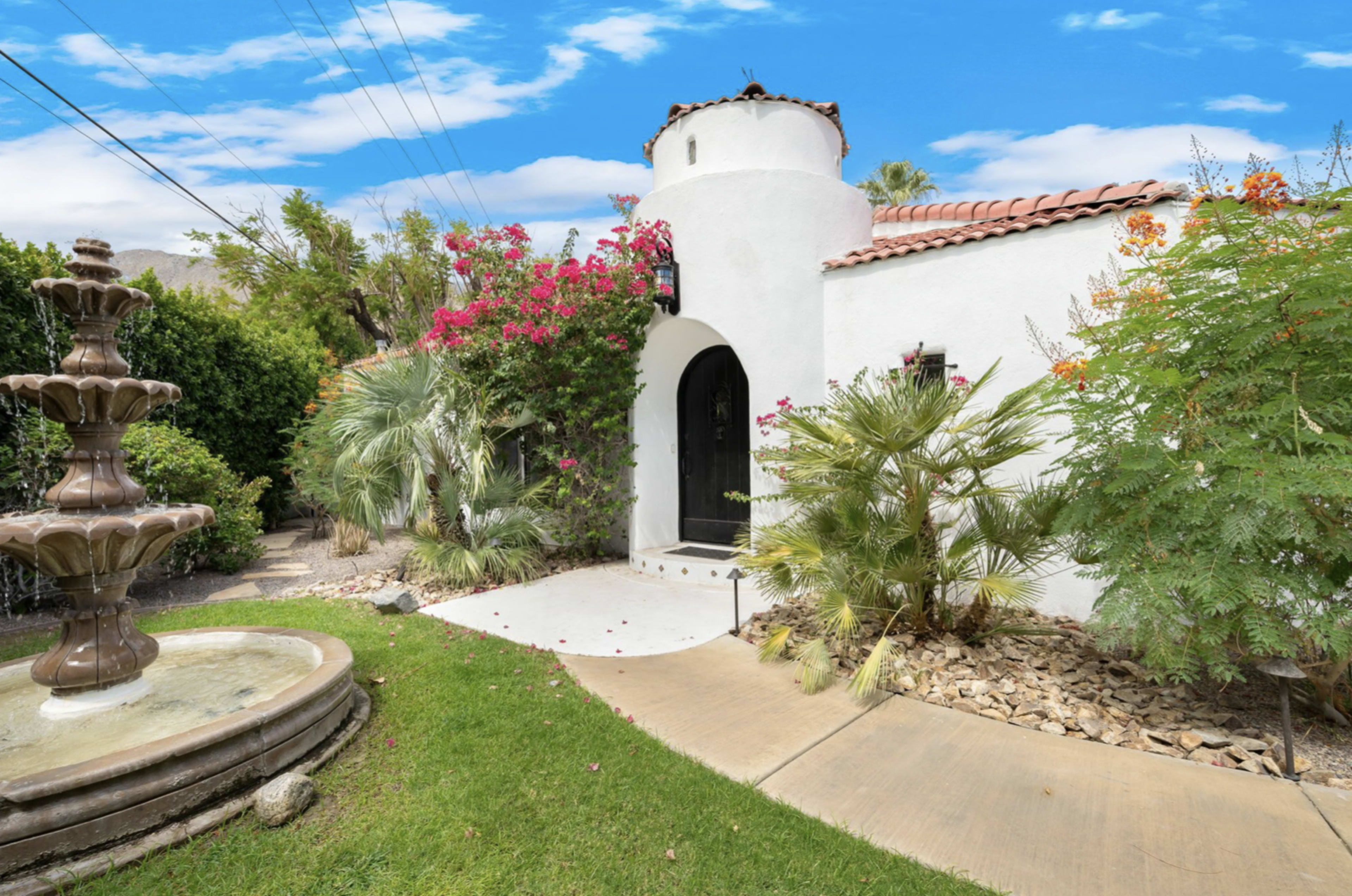 A white stucco house with a decorative entrance features a prominent fountain and colorful landscaping.