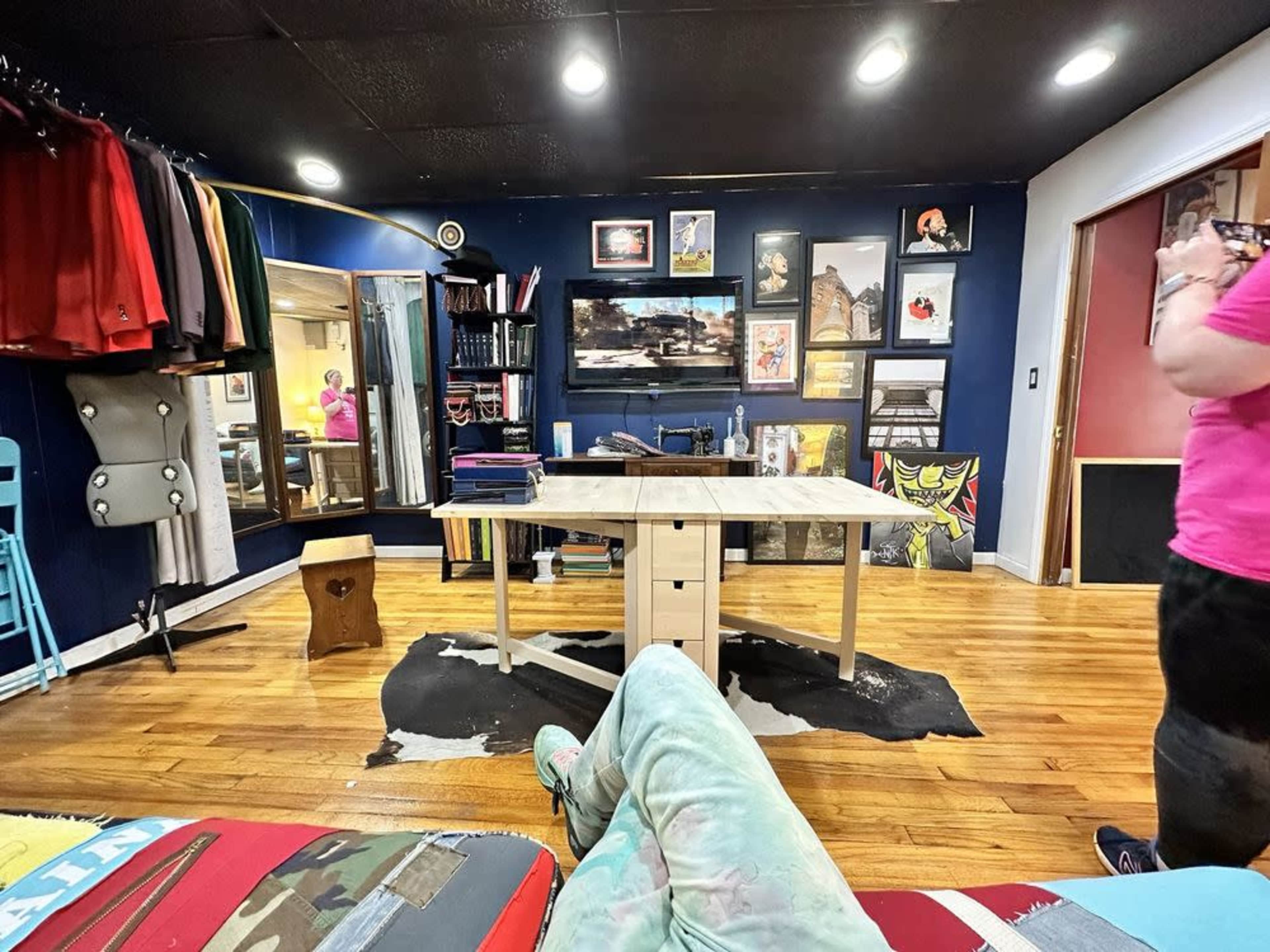 A person stands to the right in a room filled with artwork, furniture, and clothing racks, while a table is centered in the middle of the space.