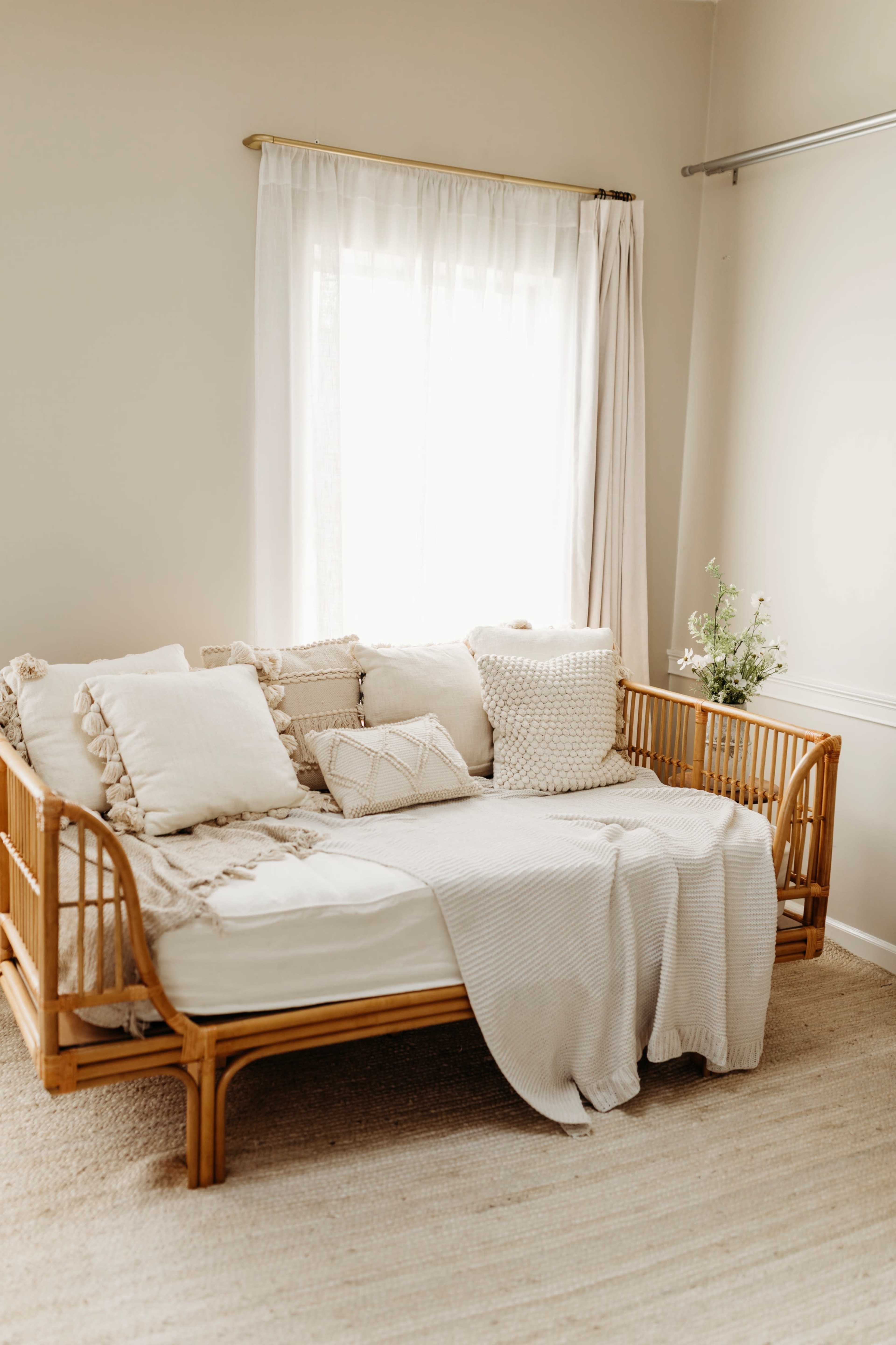 The image shows a cozy, light-colored room featuring a rattan daybed adorned with various textured pillows and a soft blanket, positioned near a window with sheer curtains.