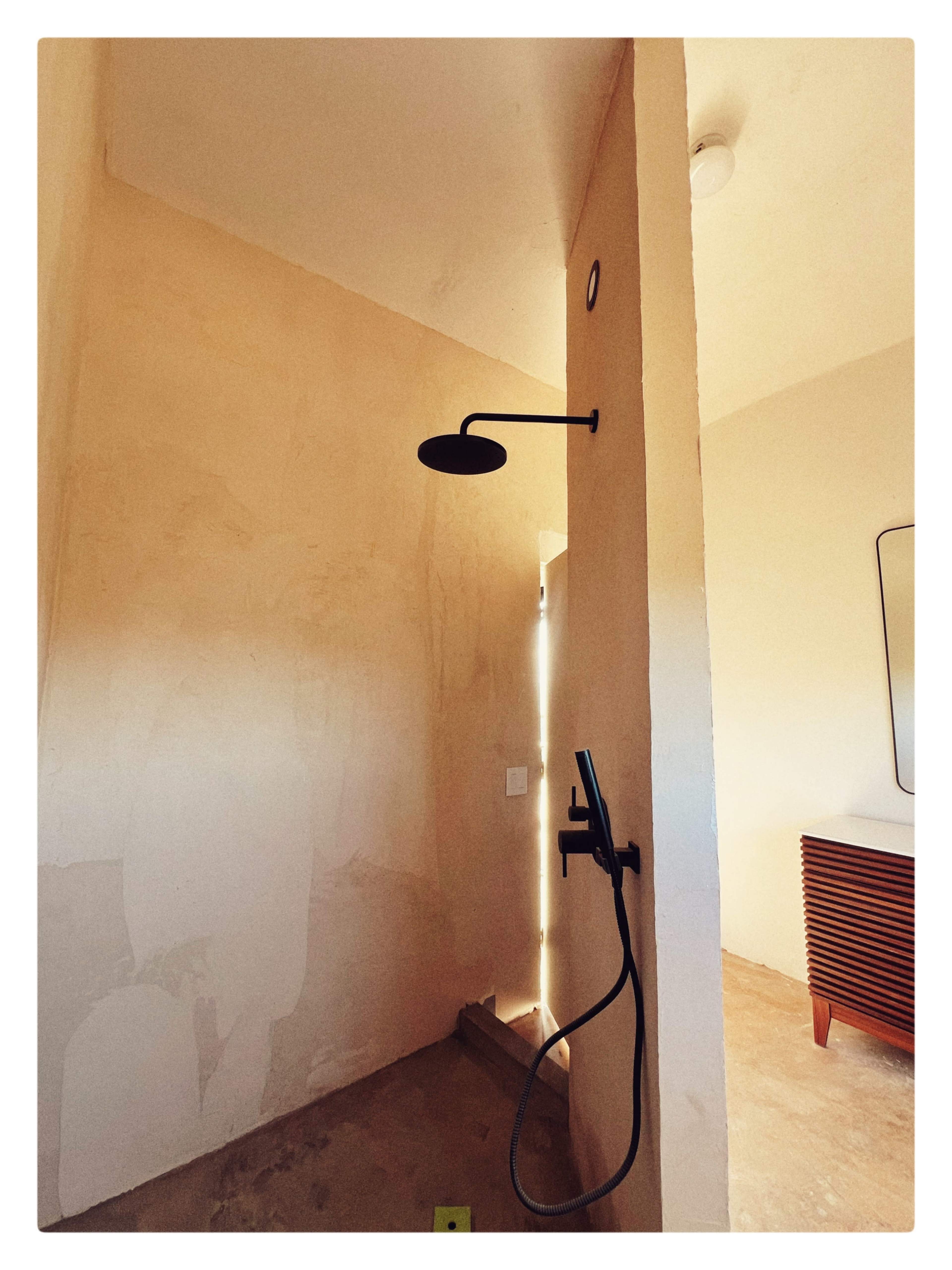 The image shows a minimalist bathroom featuring a wall-mounted showerhead and a handheld shower fixture, with a door partially open allowing light to enter.