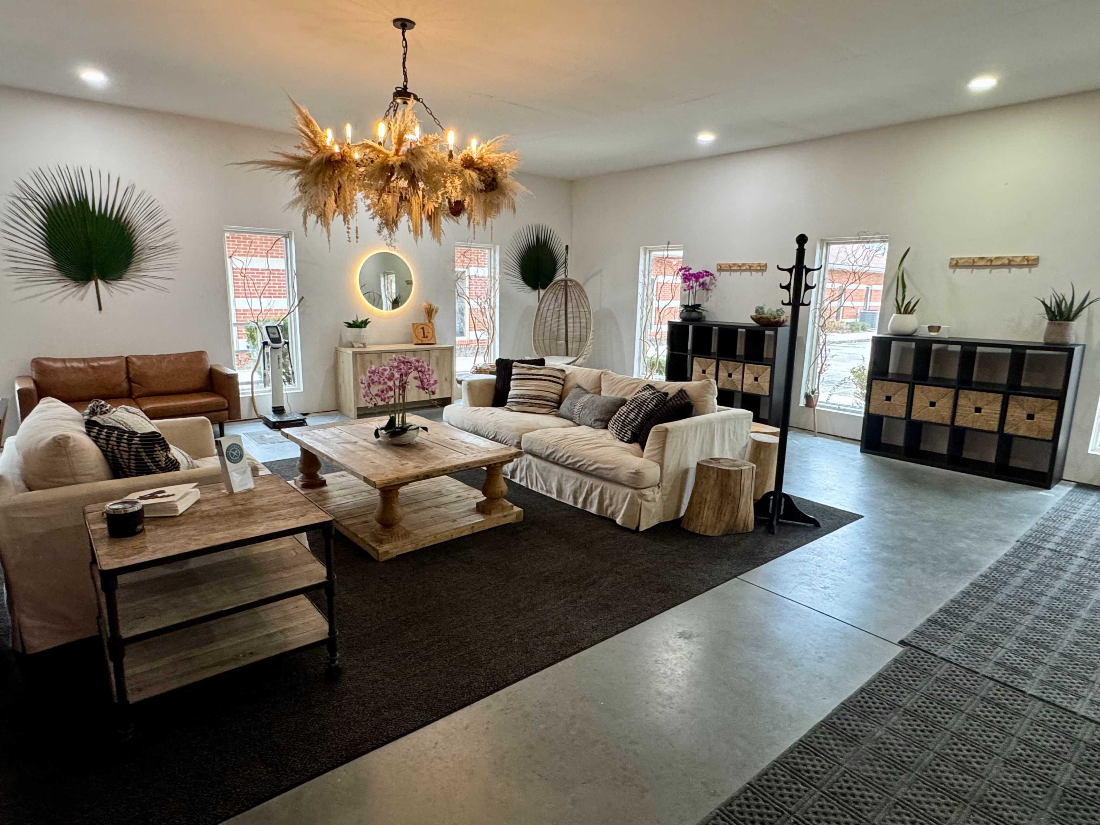 The image shows a spacious, modern living area with a light-colored sofa, a wooden coffee table, and a decorative chandelier, complemented by plants and artwork on the walls.