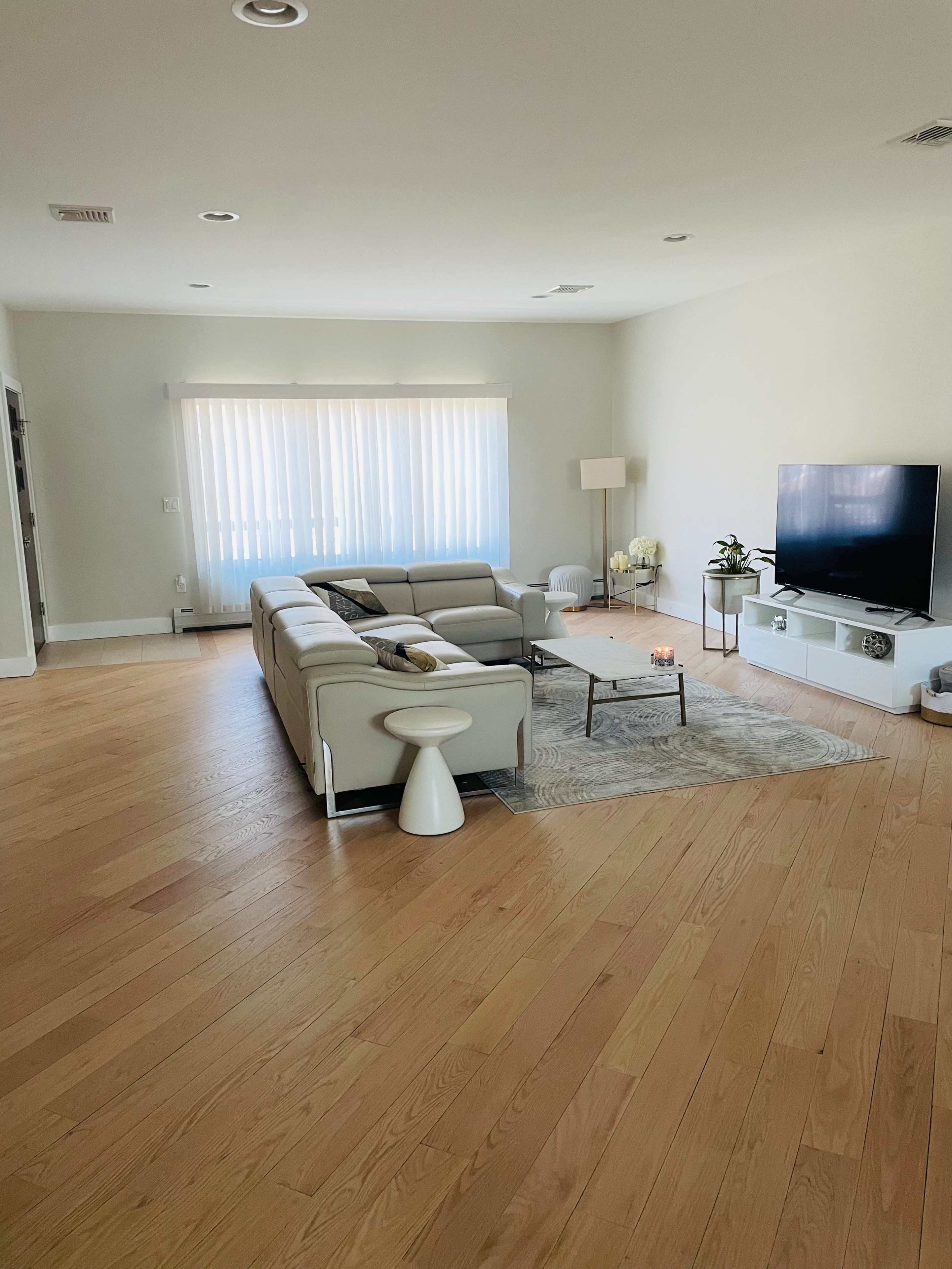 A modern living room features a light-colored sectional sofa, a coffee table, a floor lamp, and a television against a wall with large windows partially covered by sheer blinds.