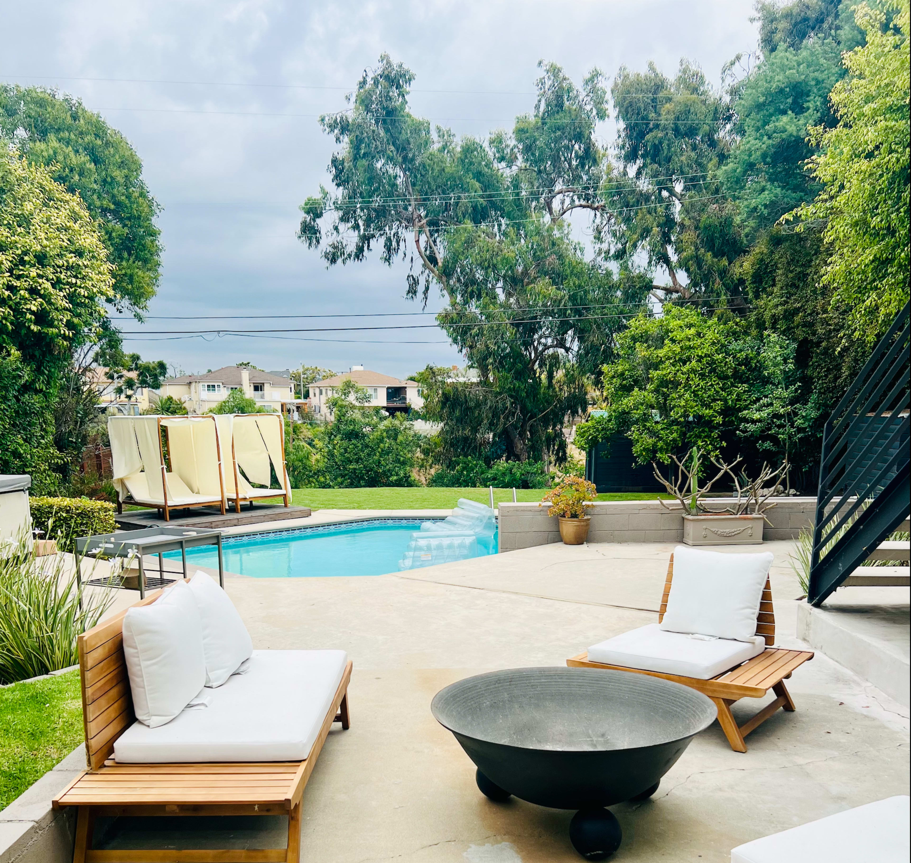 The scene features a sunny backyard with a swimming pool, two lounge chairs, a fire pit, and lush greenery in the background.
