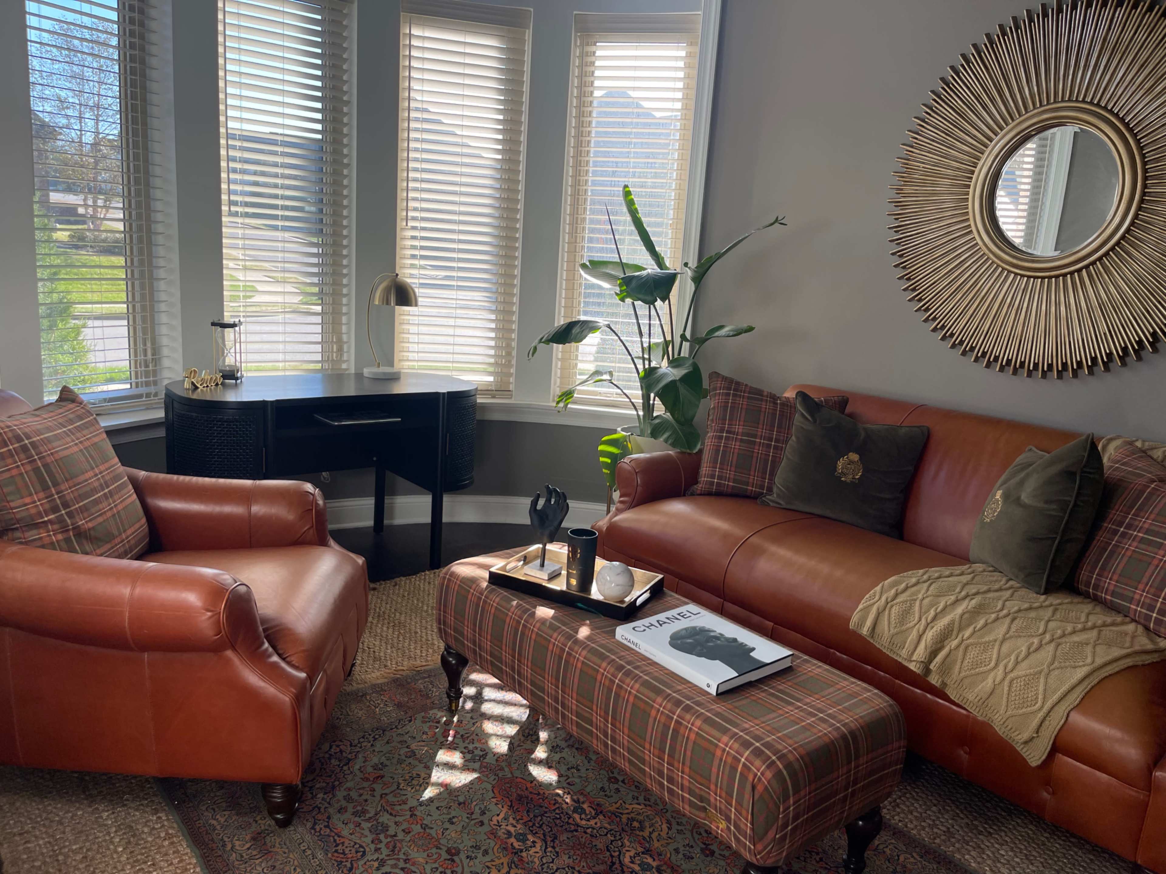 A cozy living room features a brown leather sofa and armchair, a round coffee table with books, and a desk beside large windows with blinds, surrounded by plants.