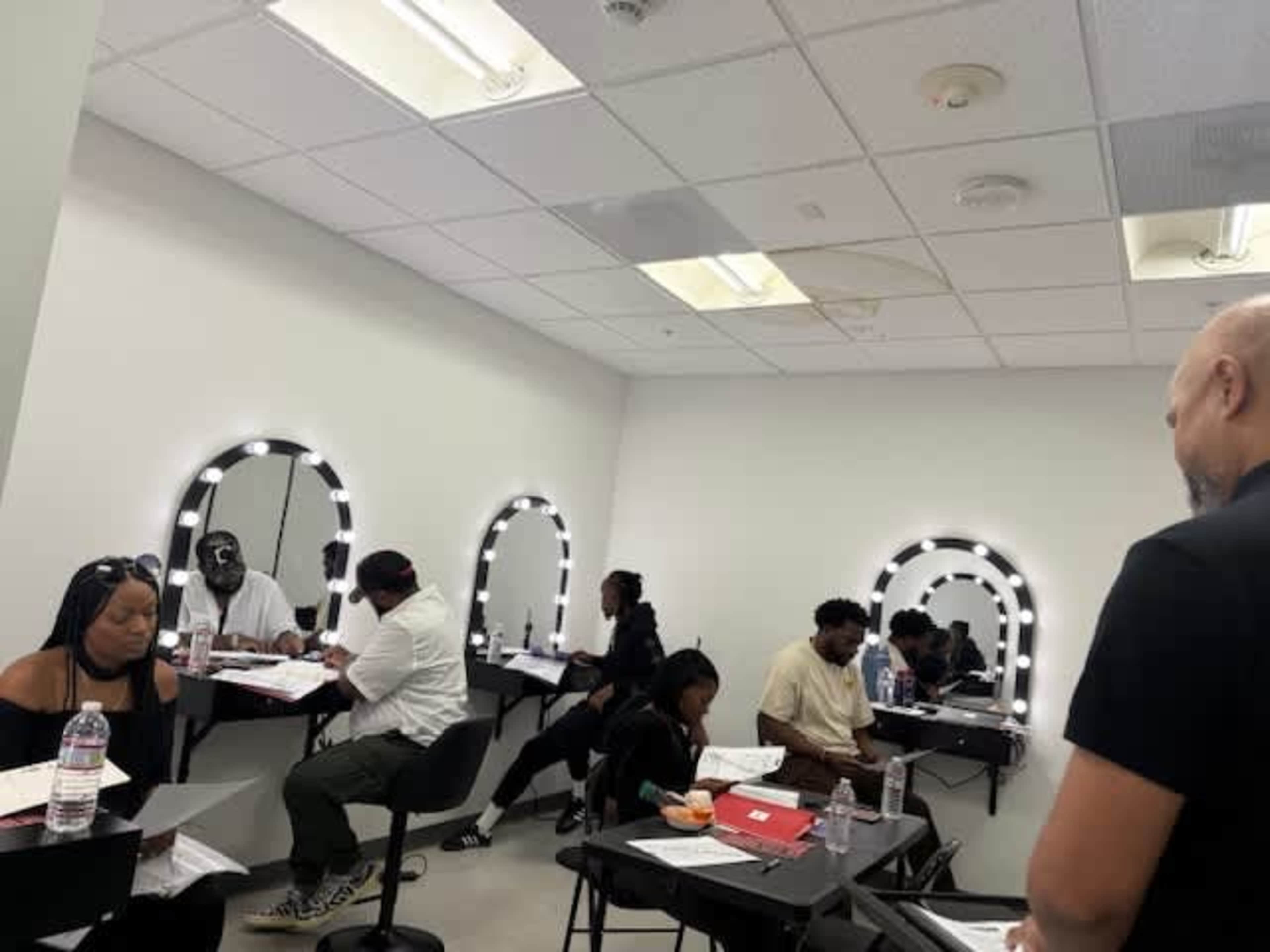 The image shows a group of people seated at tables in a well-lit room with large mirrors, engaged in a learning or collaborative session.
