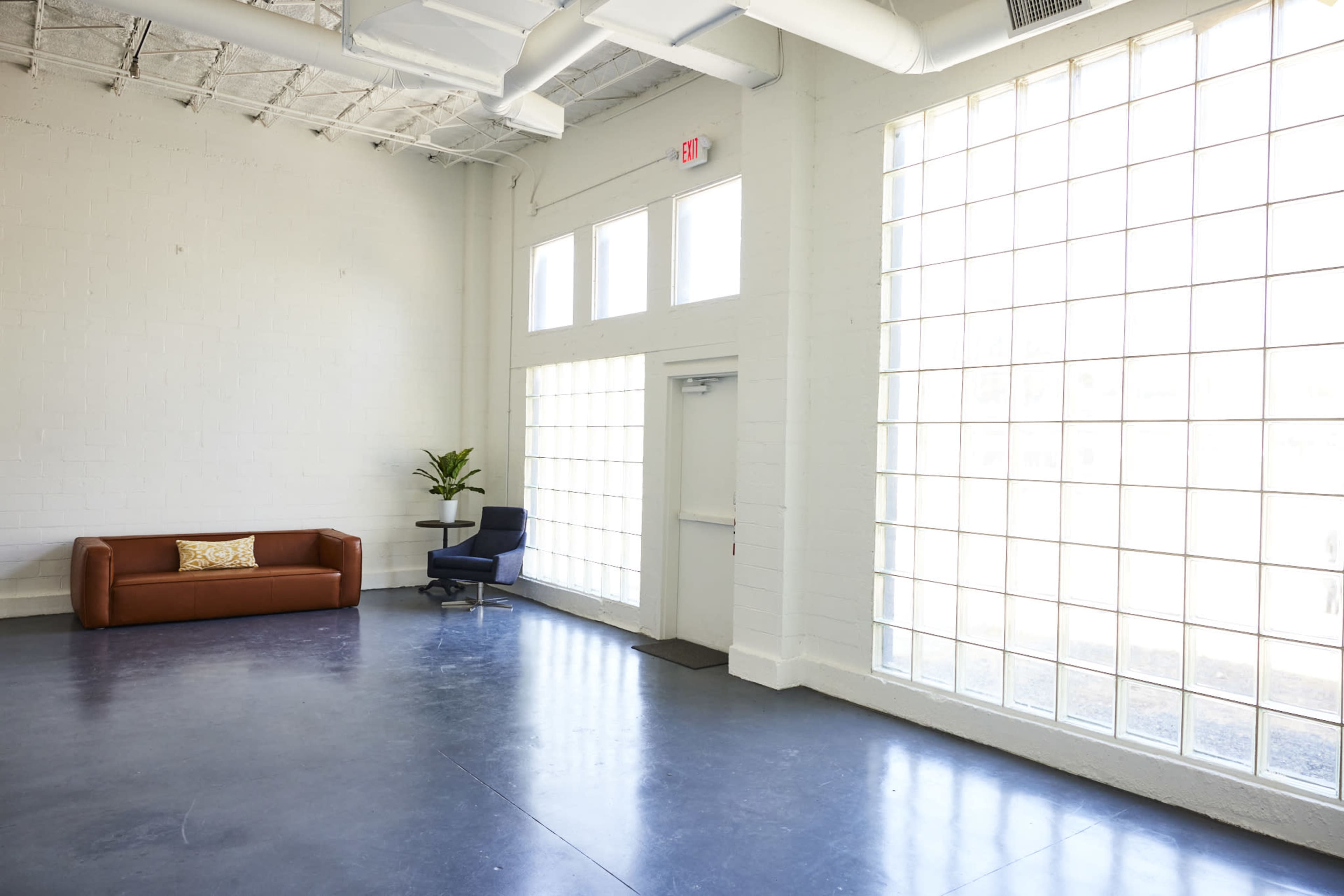 A spacious, minimally furnished room featuring a brown sofa, a blue chair, and large windows allowing ample natural light.