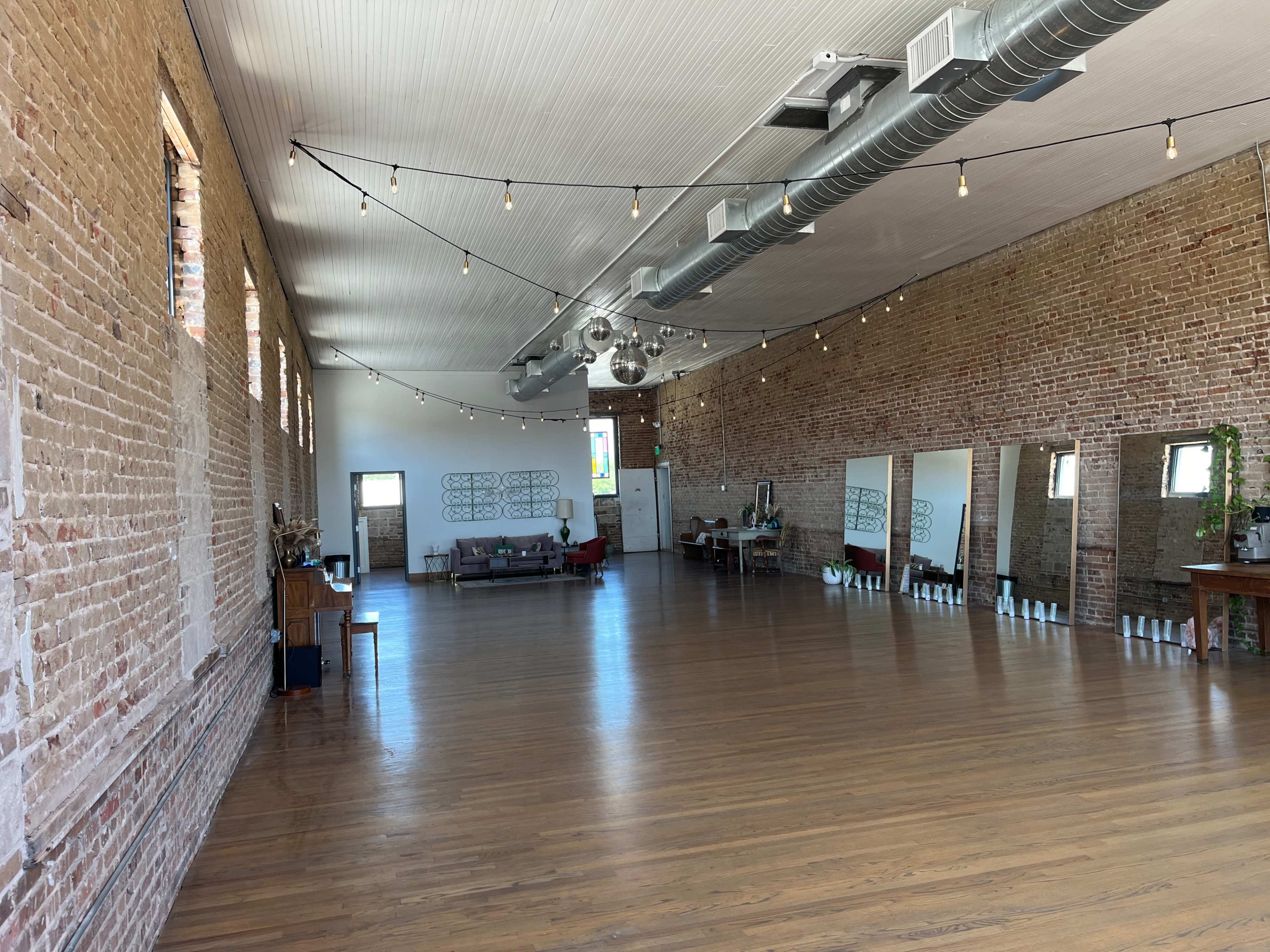 A spacious room with exposed brick walls, wooden flooring, and several mirrors along one side, illuminated by string lights.