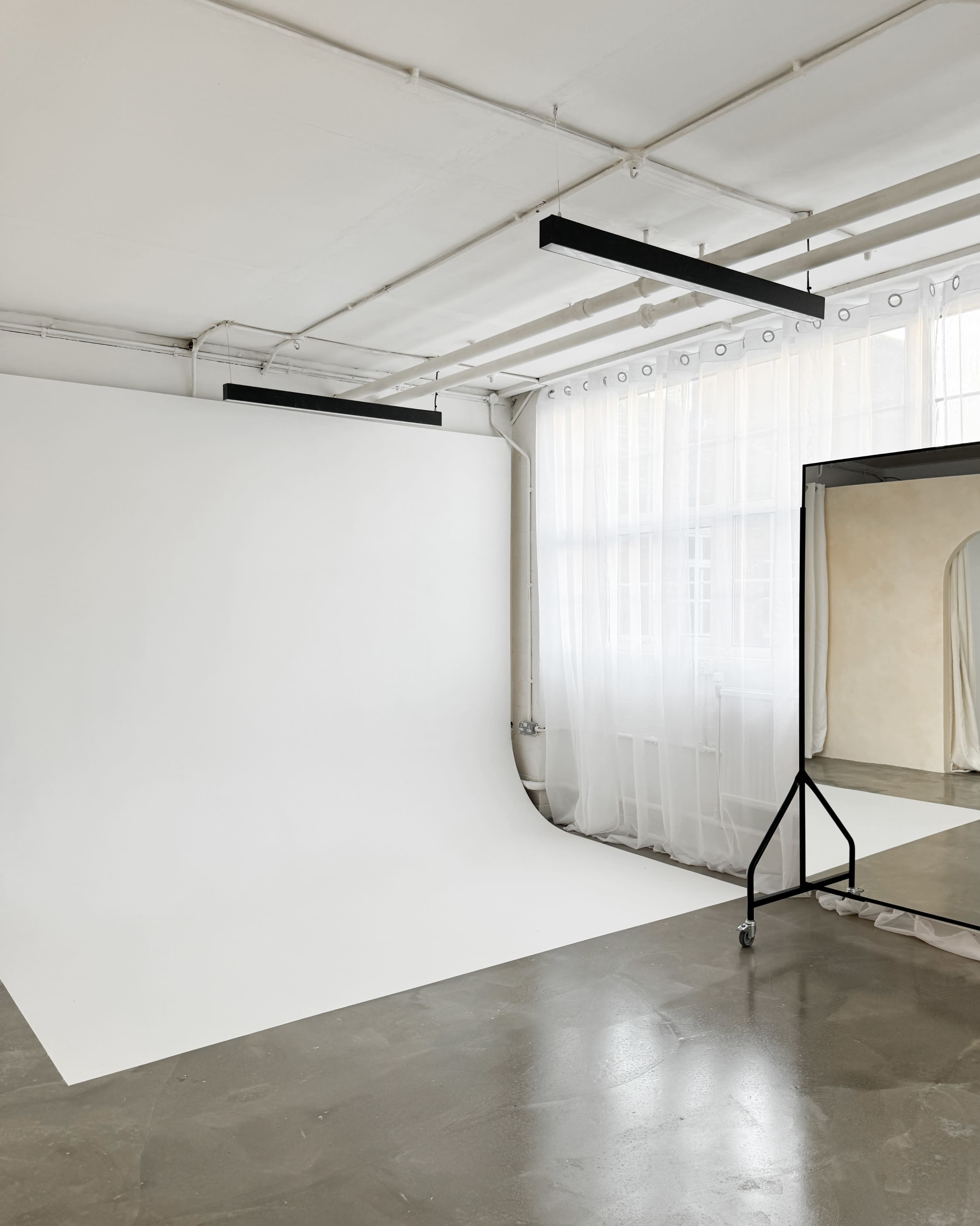 The image shows a spacious studio with a seamless white backdrop, large windows with sheer curtains, and a full-length mirror on a stand.
