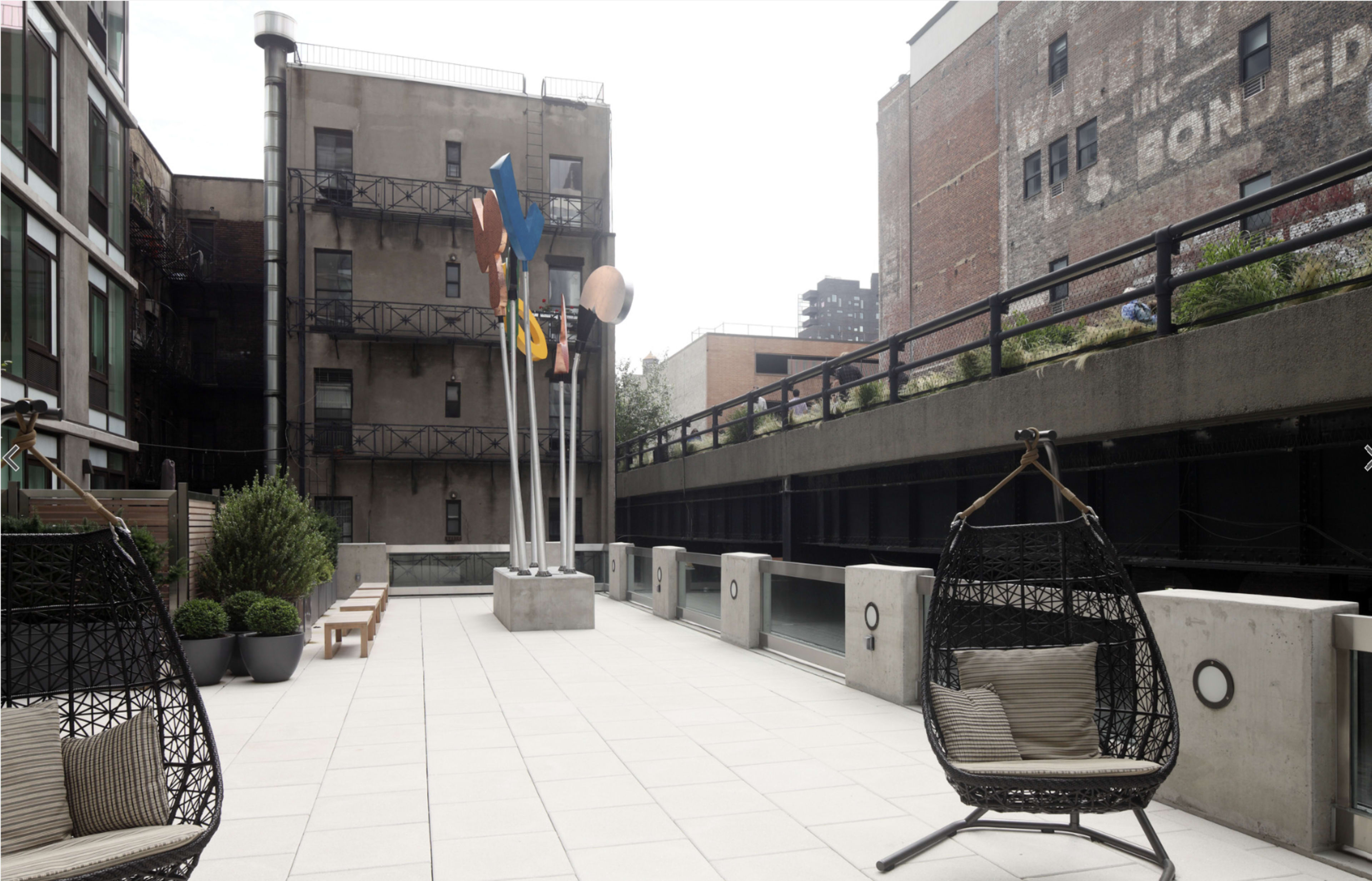 The image shows a modern rooftop patio featuring hanging chairs, potted plants, and several colorful sculptures near a building with industrial architecture.