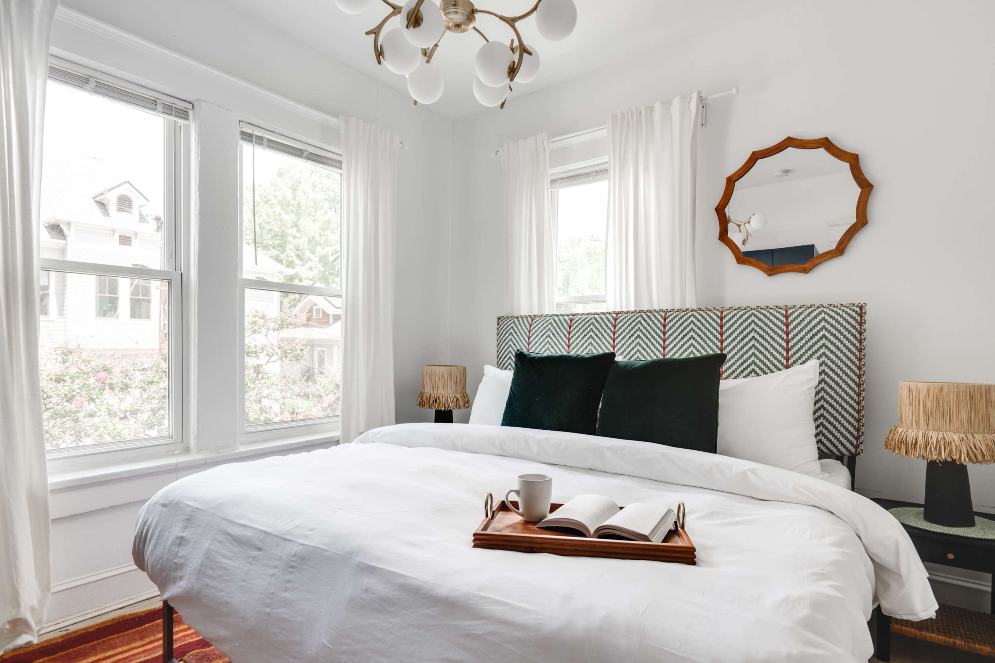 A neatly arranged bedroom features a bed with a white duvet, green accent pillows, and a wooden tray holding a book and a cup, beside large windows adorned with sheer curtains.