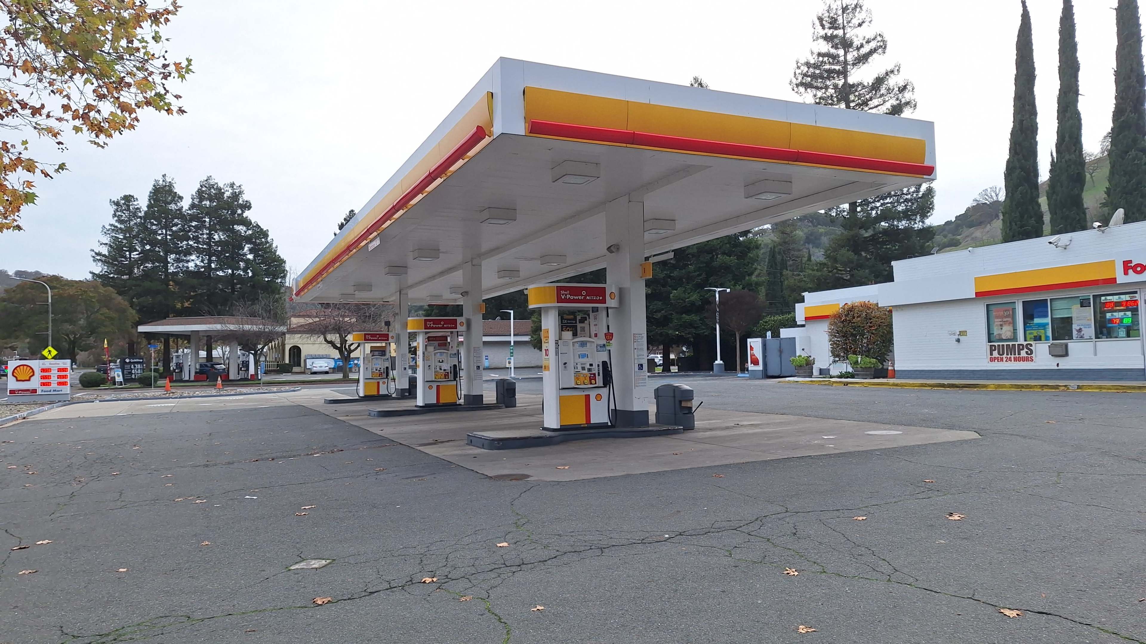 An empty gas station with several fuel pumps under a large canopy, surrounded by trees and a convenience store nearby.