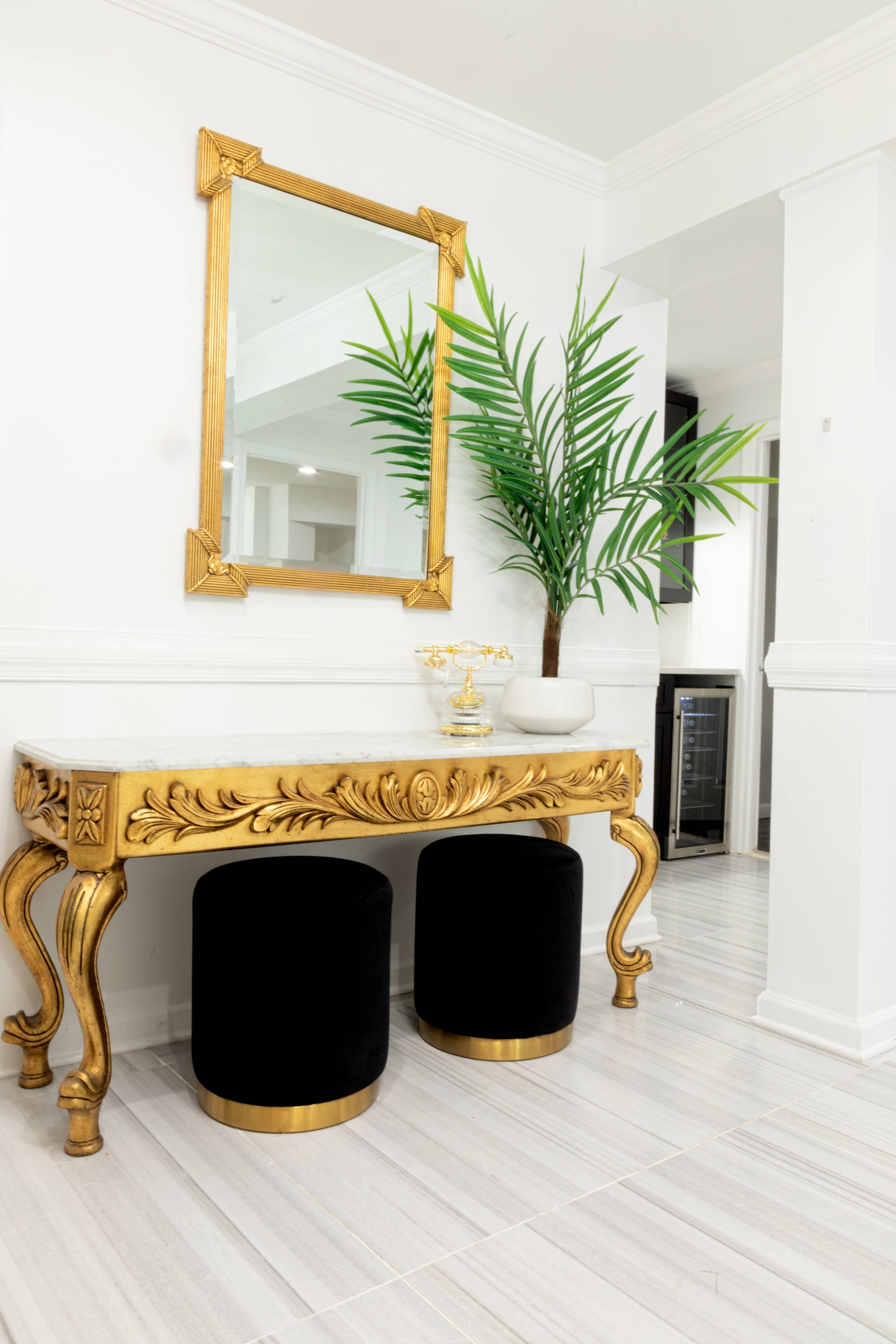 A gold-accented console table stands against a white wall, featuring a large mirror and a potted palm, accompanied by two black upholstered stools.