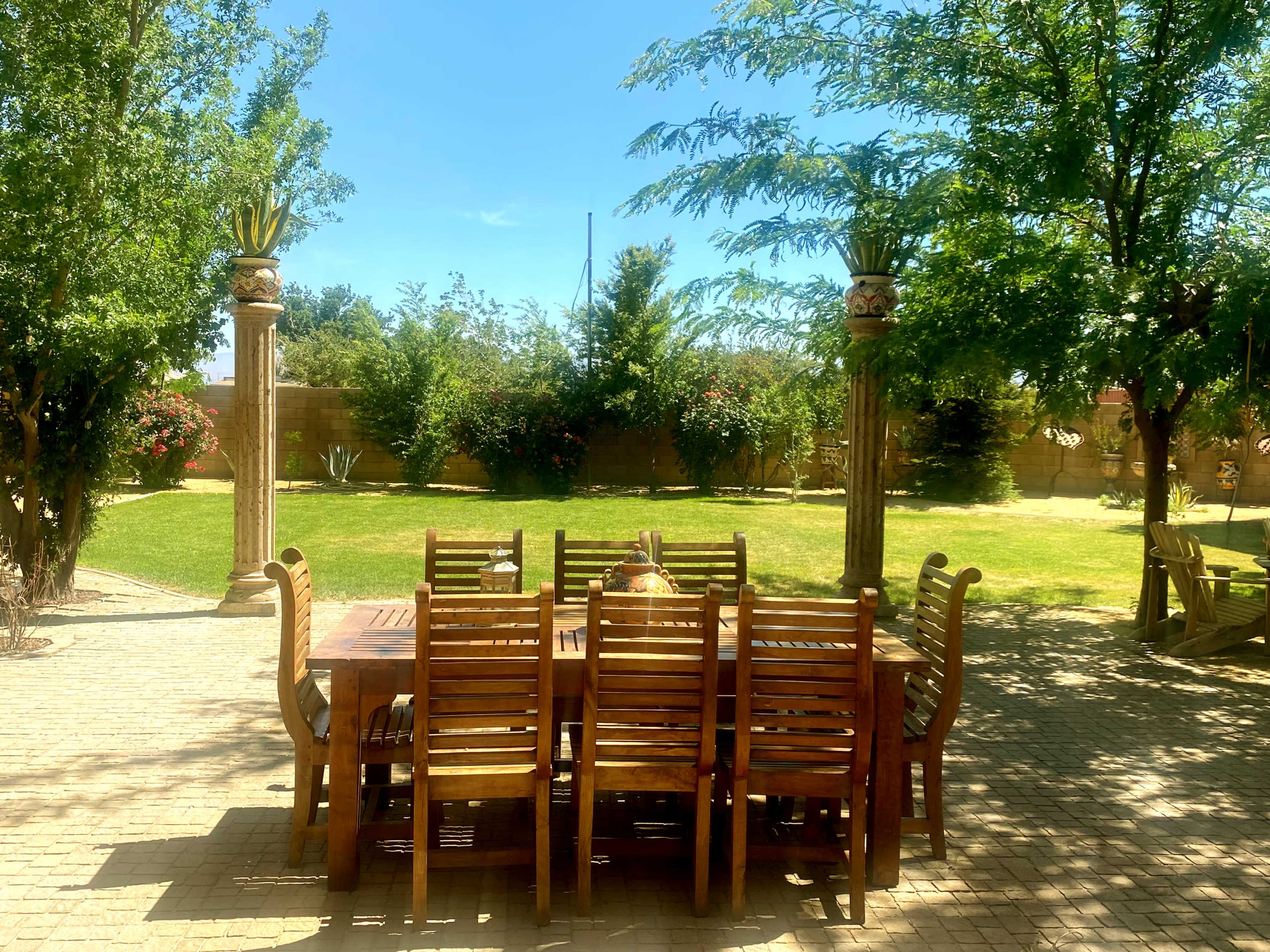 A wooden dining table with multiple chairs is set in a spacious garden surrounded by greenery and trees.