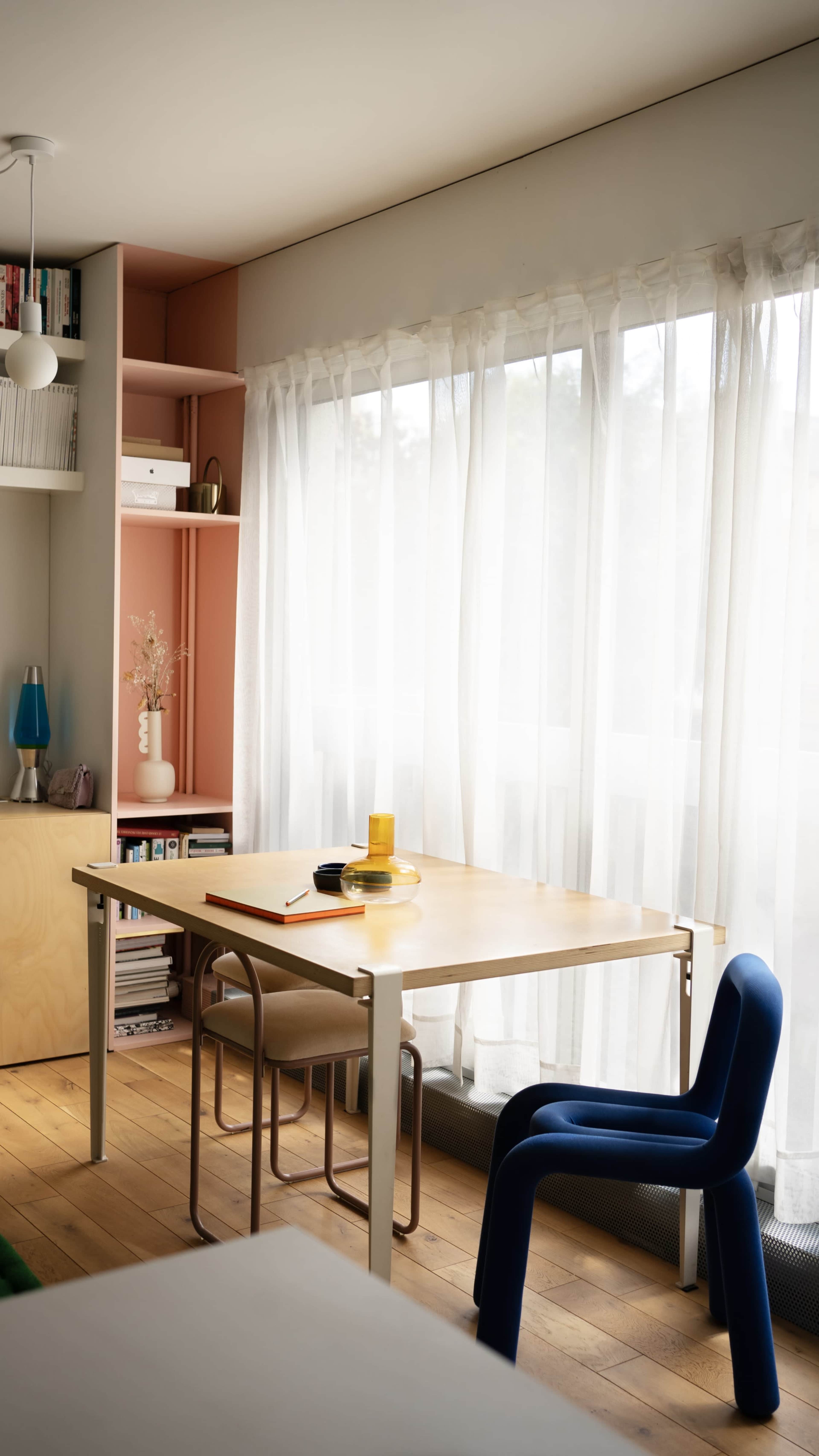 A modern room features a wooden table with a round black plate and a yellow glass, complemented by a blue chair and natural light filtering through sheer curtains.