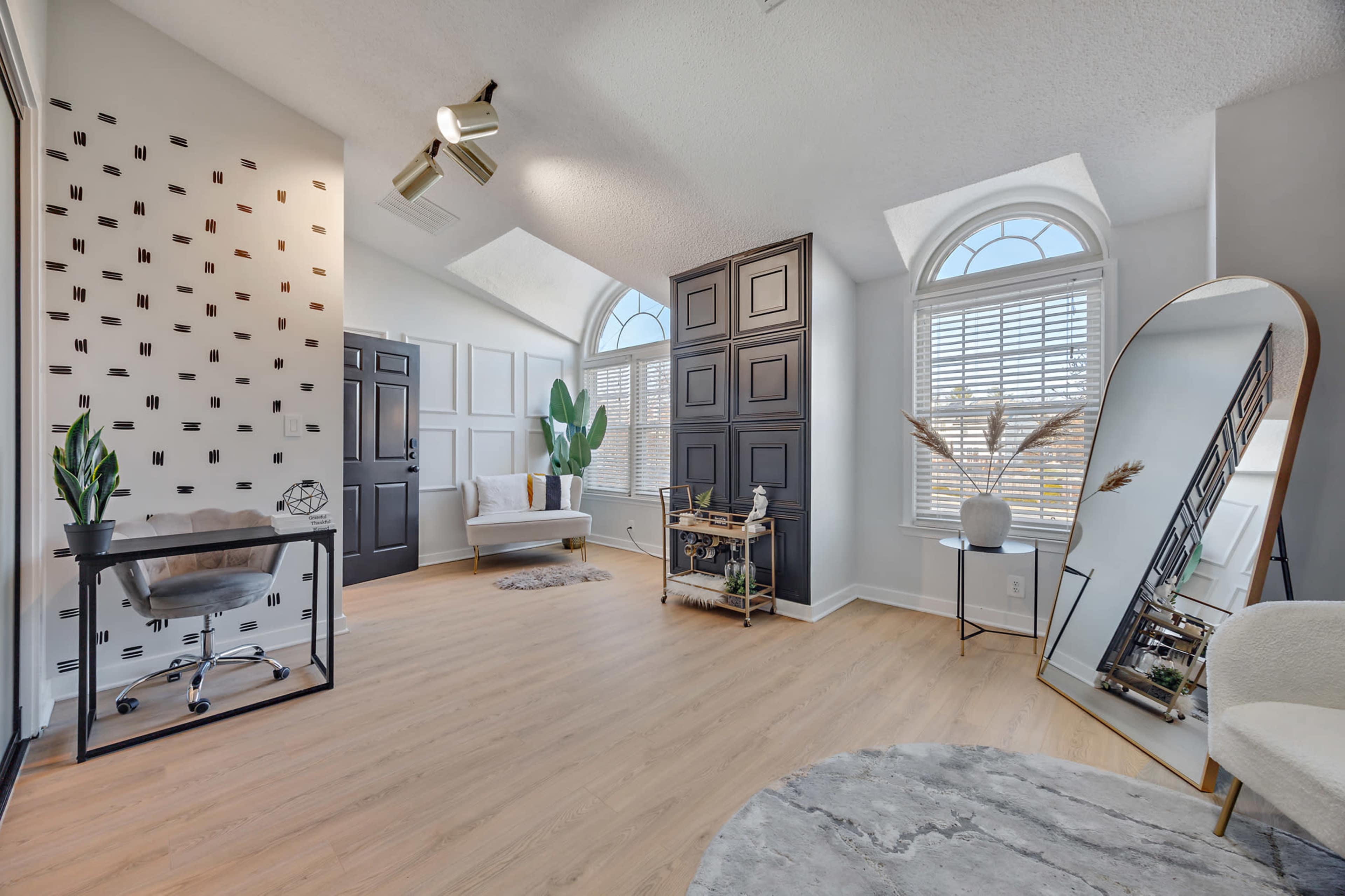 The image shows a bright, contemporary interior space featuring wooden flooring, large windows, and modern furnishings, including a desk and a mirror.