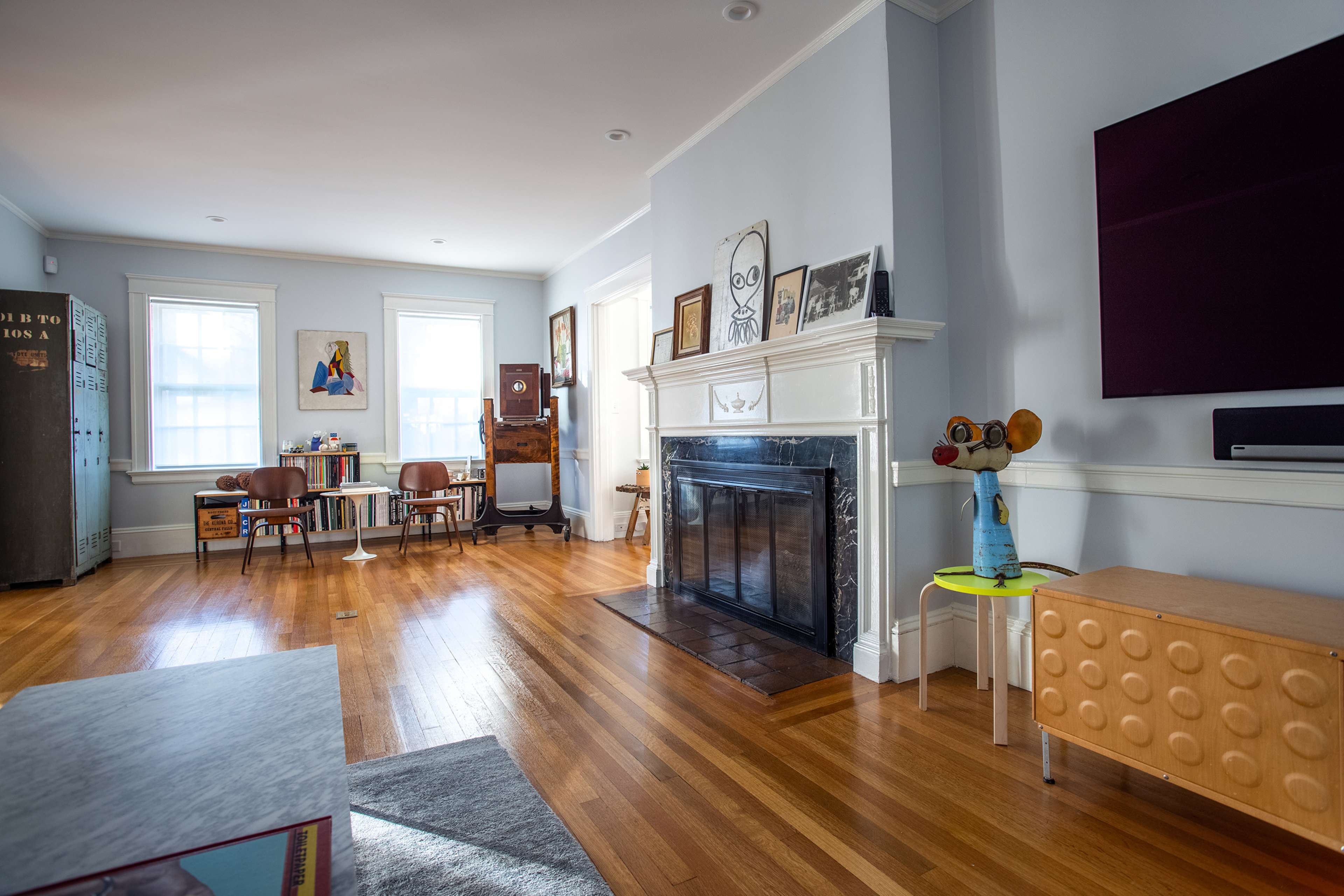A bright living room with hardwood floors, a fireplace, and various furniture pieces, including a colorful side table and a wall-mounted television.