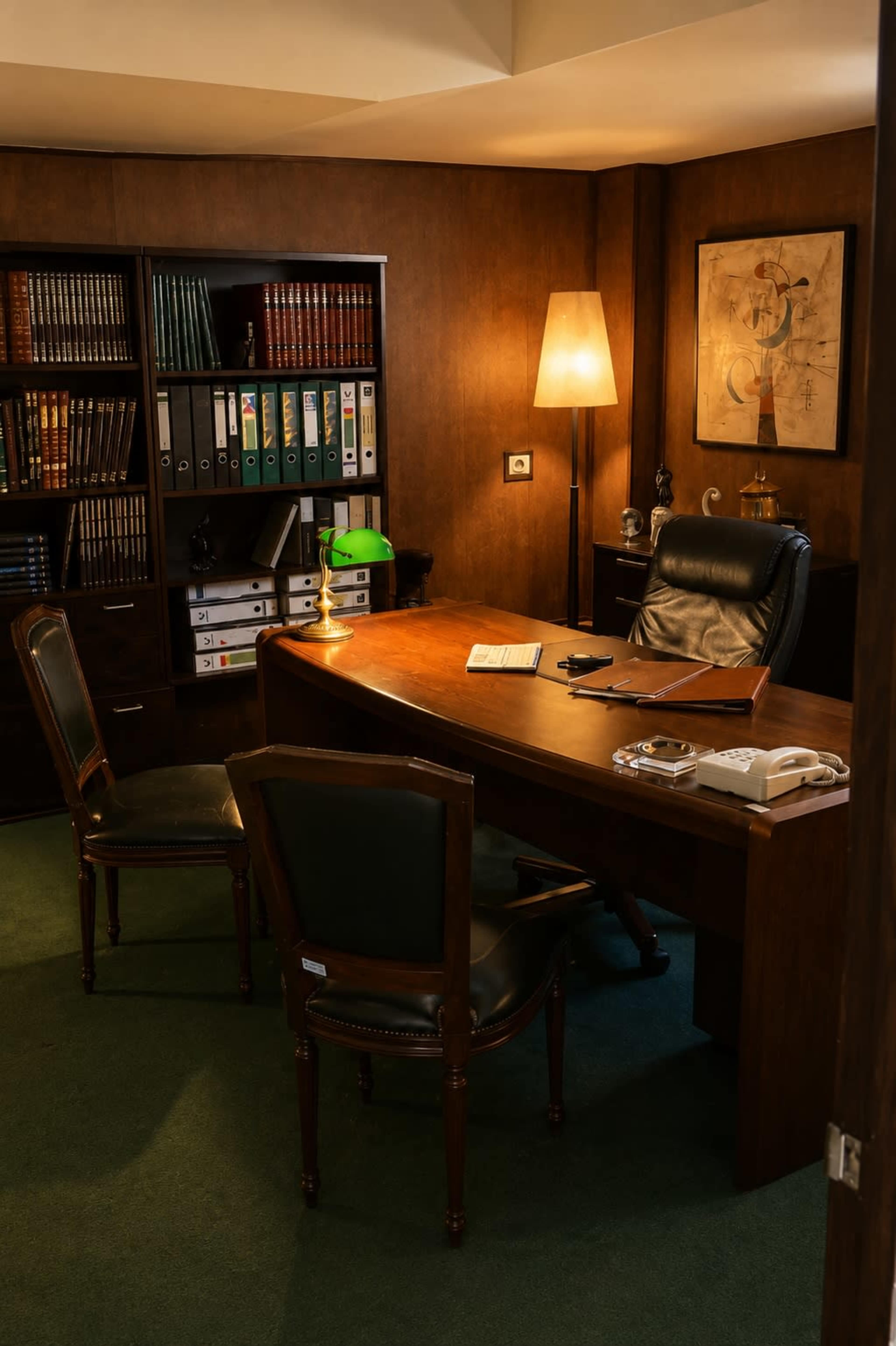 The image shows a dimly lit office with a wooden desk, two chairs, a lamp, and a bookshelf filled with books and binders.