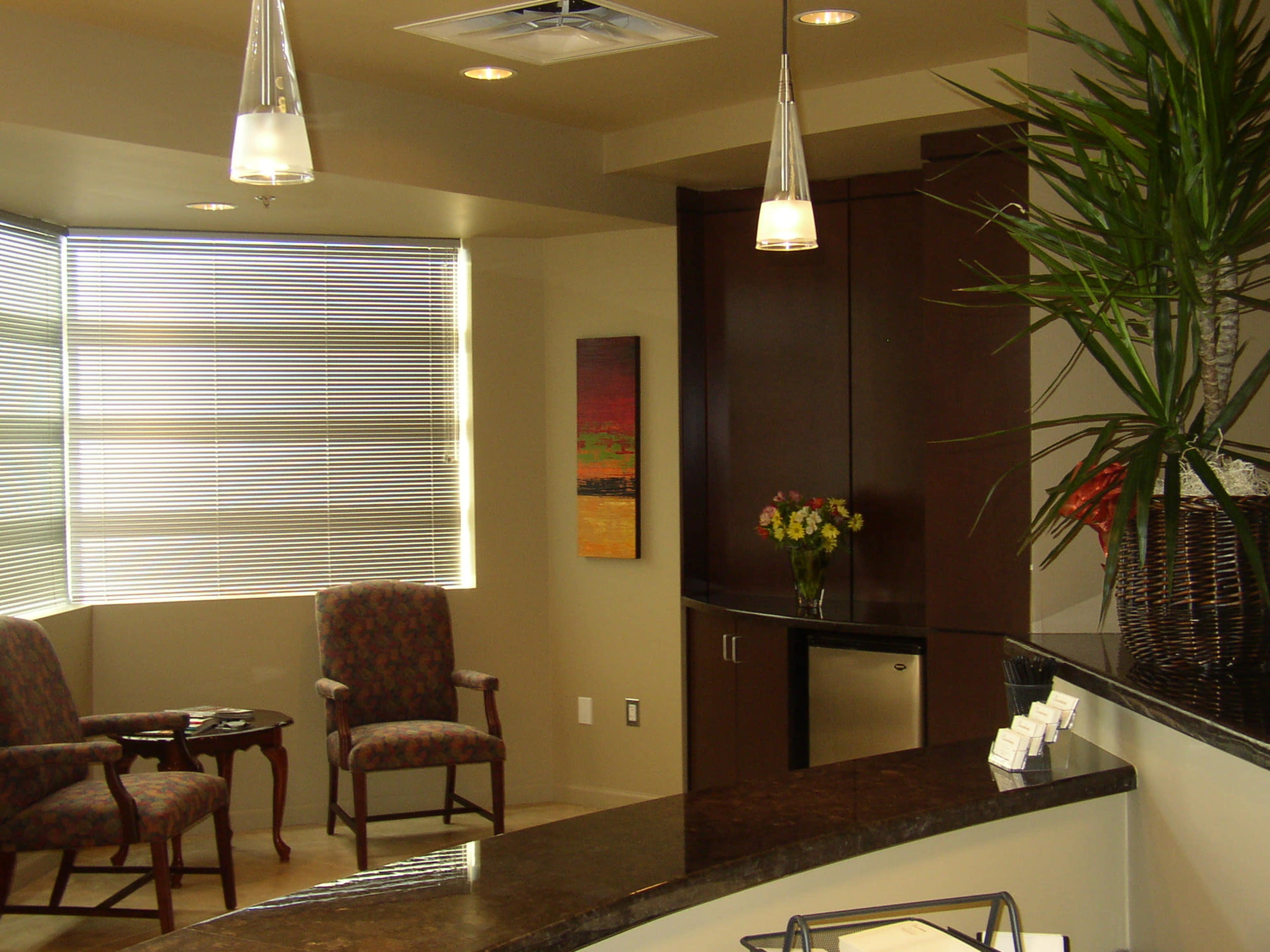 A reception area with two chairs, a small table, and a kitchenette featuring dark cabinetry and a decorative plant in the corner.