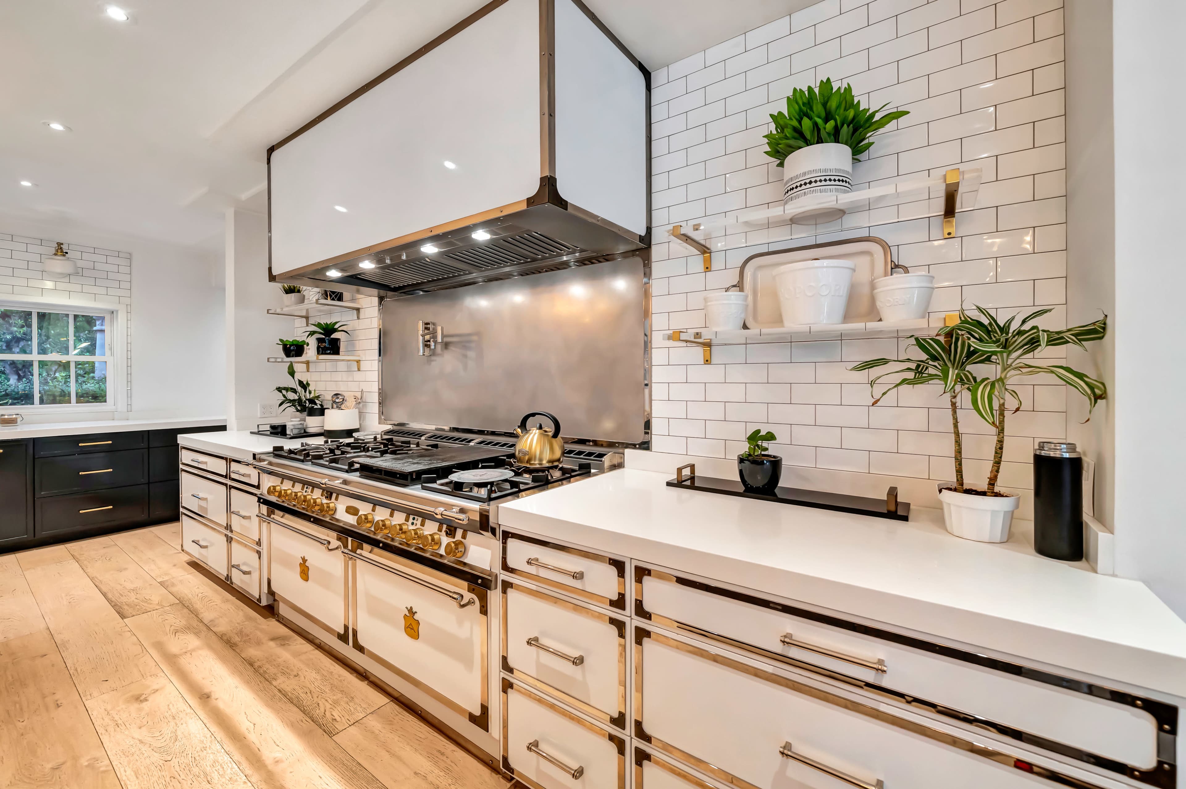 The image shows a modern kitchen with a large stainless steel range hood, an organized countertop, and white cabinets accented by black shelves and greenery.