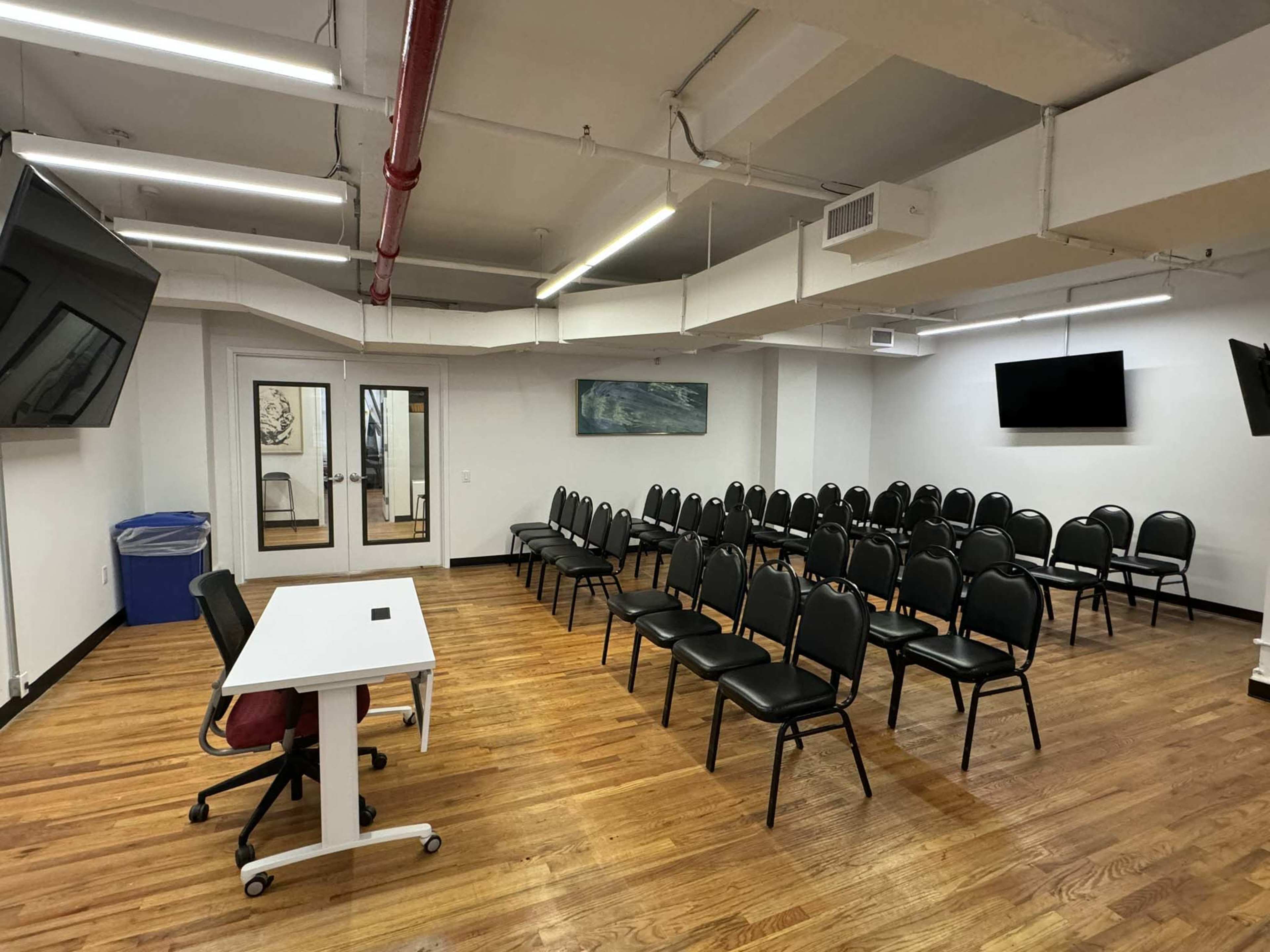 A training room features rows of black chairs facing a white table with a microphone, set against a backdrop of wooden flooring and two large screens mounted on the walls.