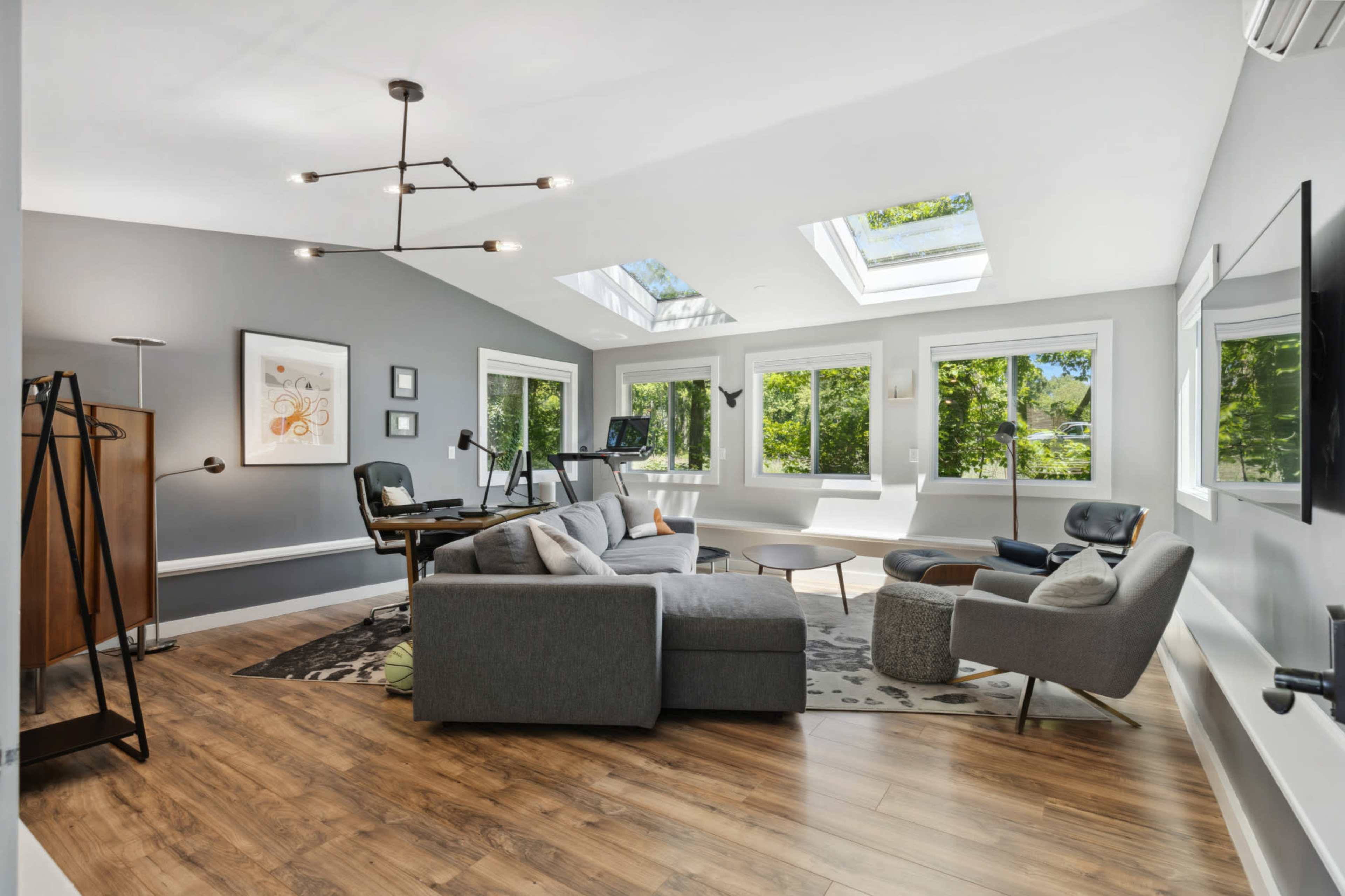 The image shows a modern living room with a sectional sofa, several windows allowing natural light, and hardwood flooring.
