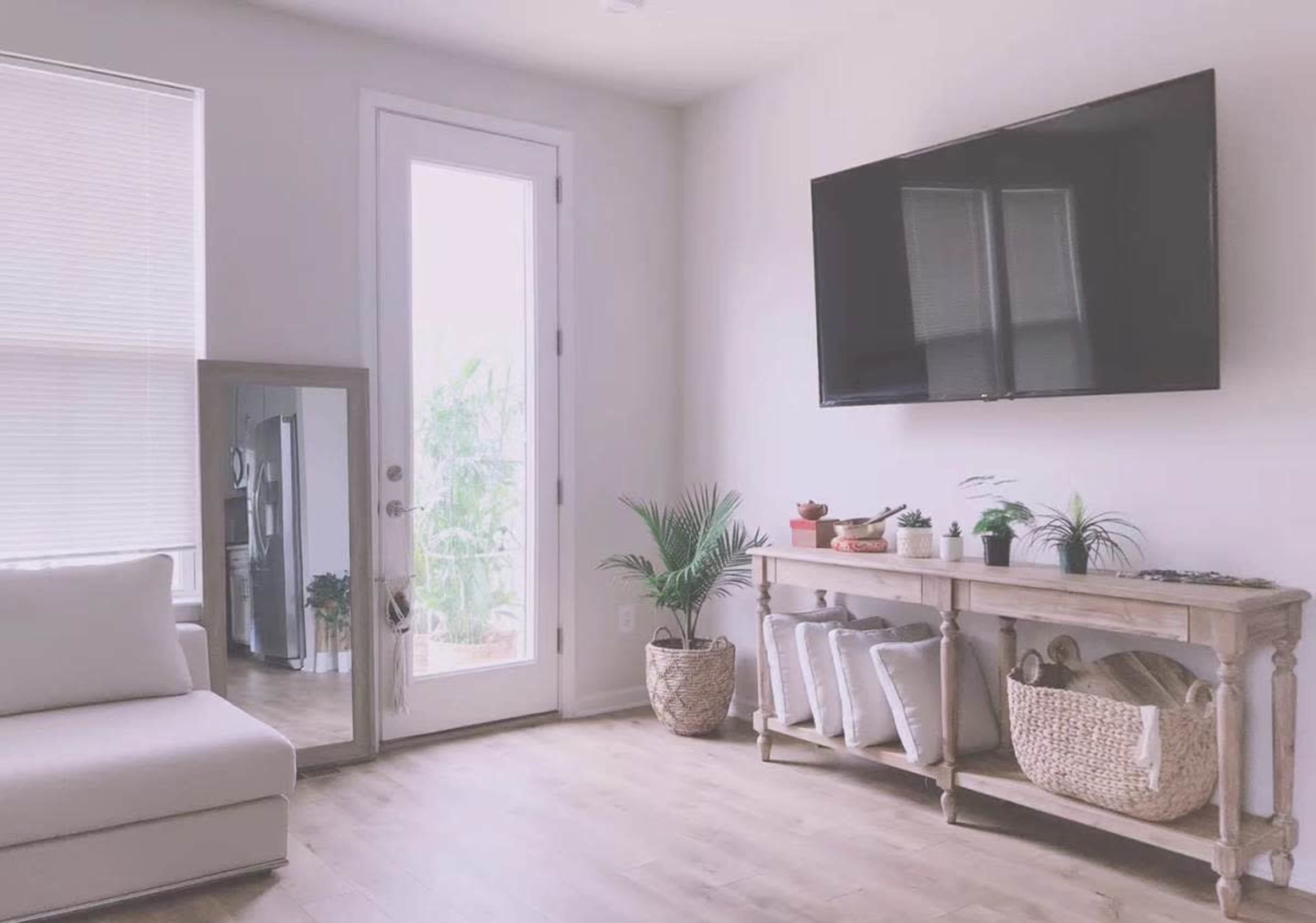 A modern living room features a gray sofa, a large wall-mounted TV, a wooden console with plants, and a glass door leading outside.