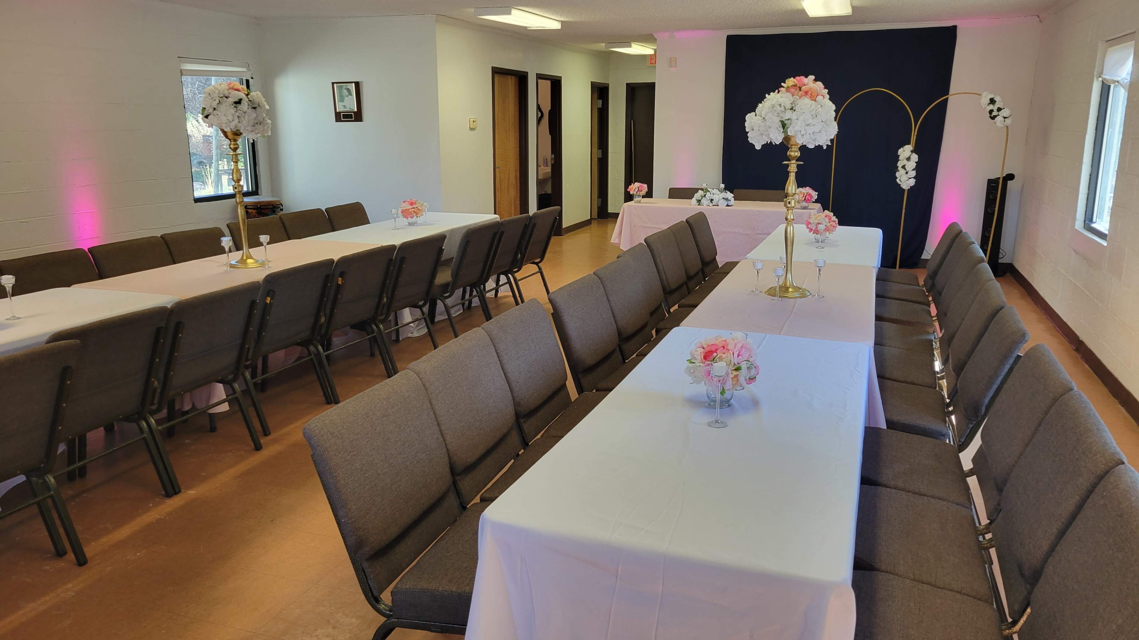 The image shows a large room set up for an event, featuring long tables covered with white tablecloths, surrounded by chairs, and decorated with floral centerpieces.