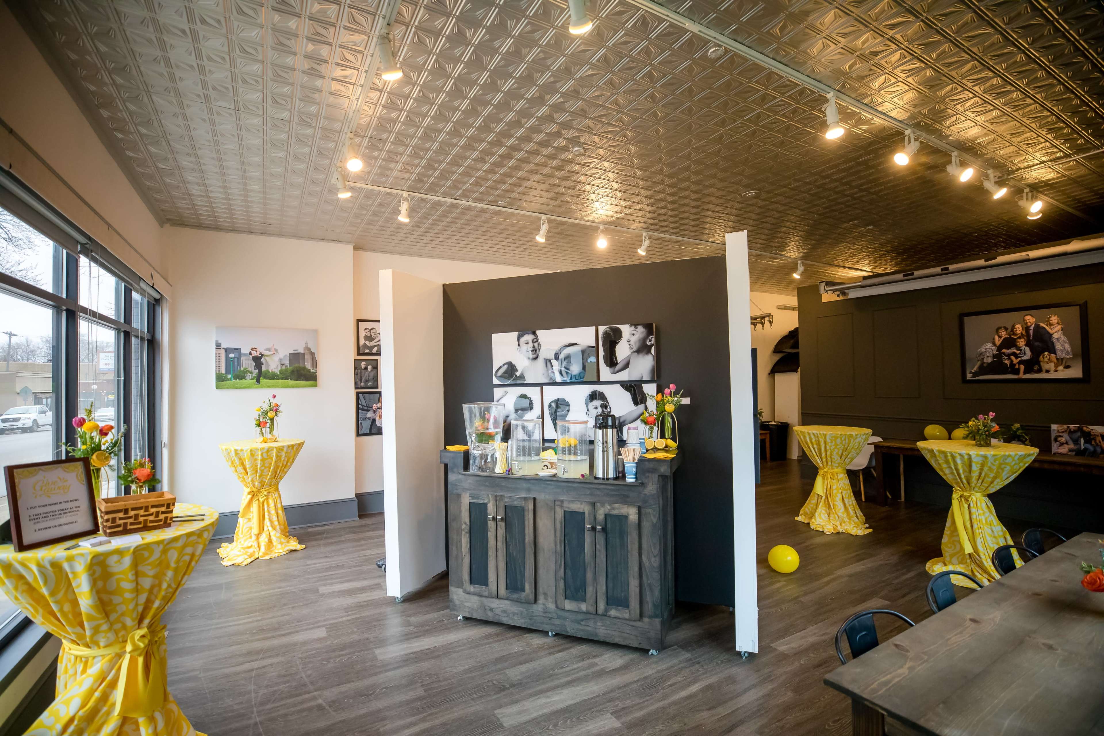 The interior of a brightly lit event space features a serving station against a black wall, surrounded by decorated tables with yellow tablecloths.