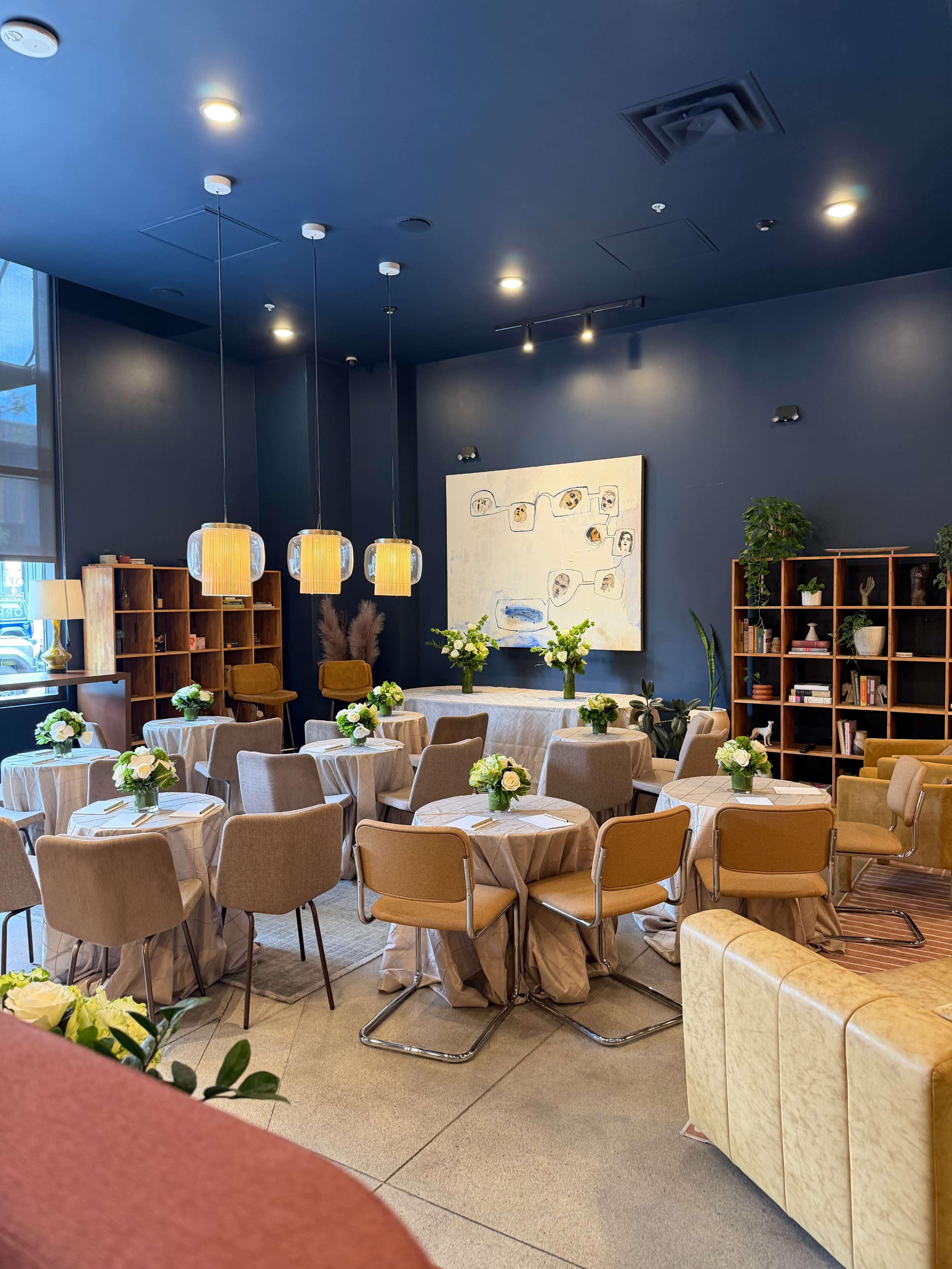 The image shows a room with several round tables set for a gathering, featuring flower arrangements and soft lighting from hanging pendant lights.