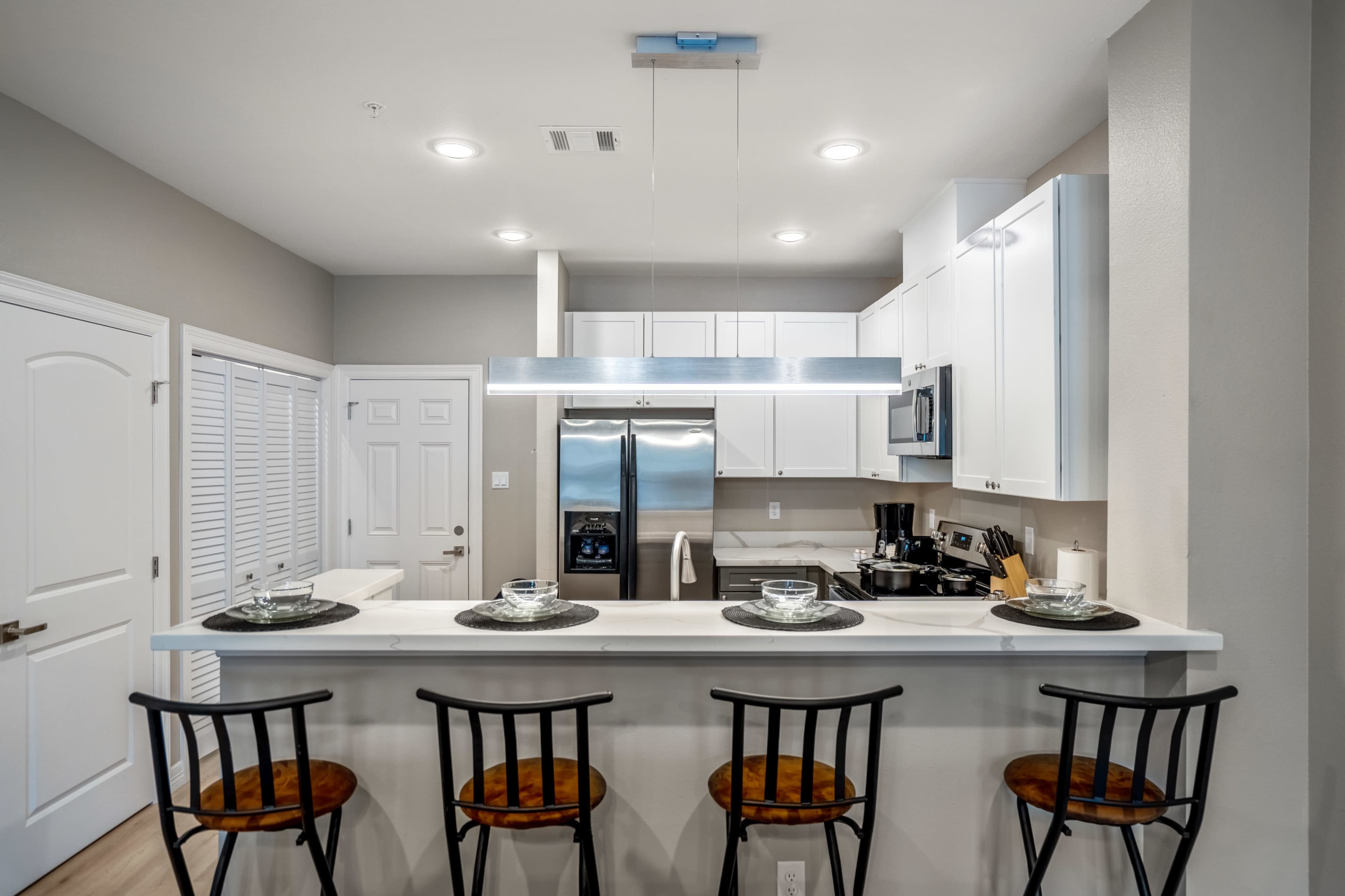 A modern kitchen features white cabinetry, stainless steel appliances, and a bar with four black chairs.