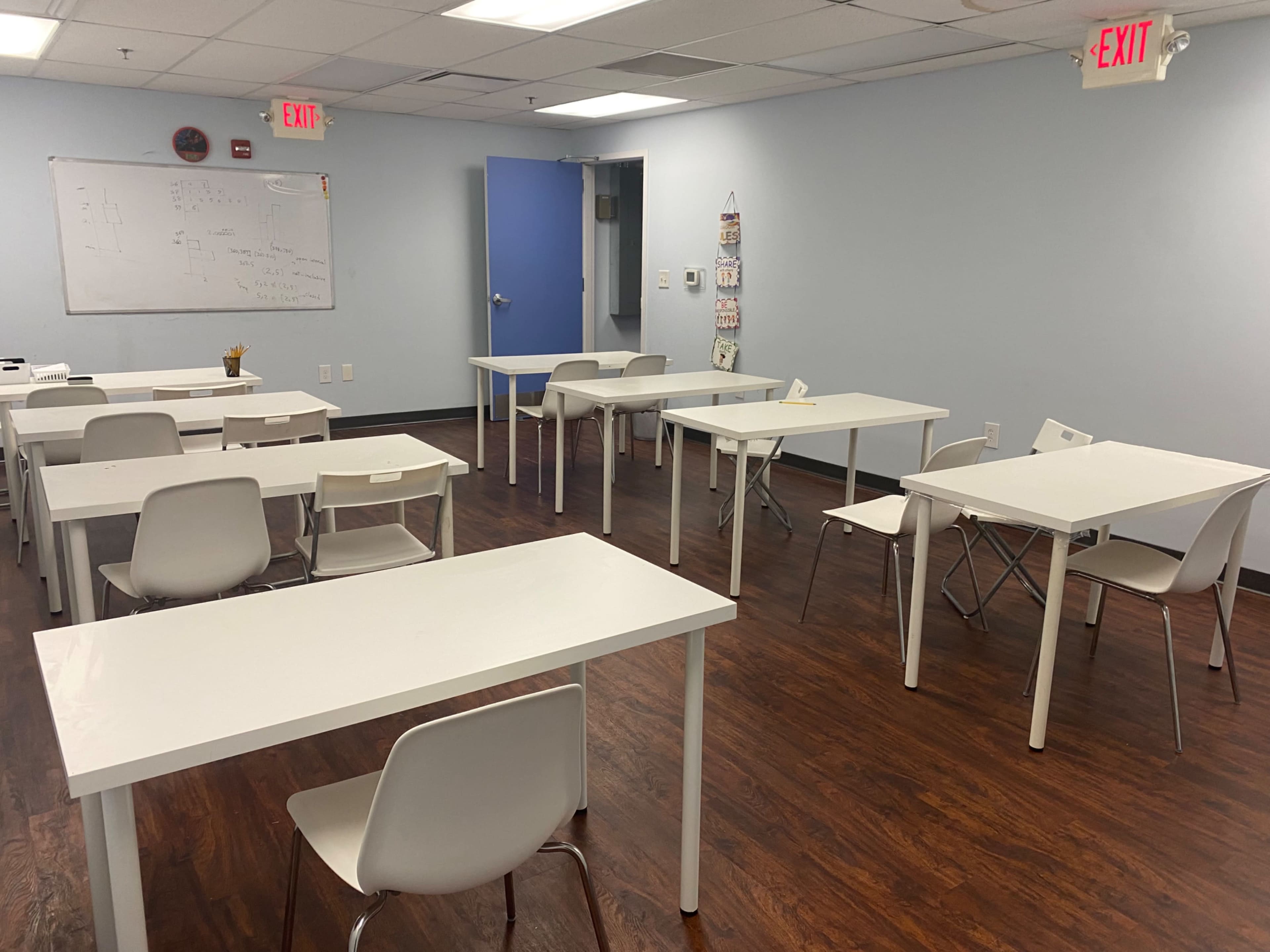 A classroom with several white tables and chairs arranged in a spacious, light blue room.