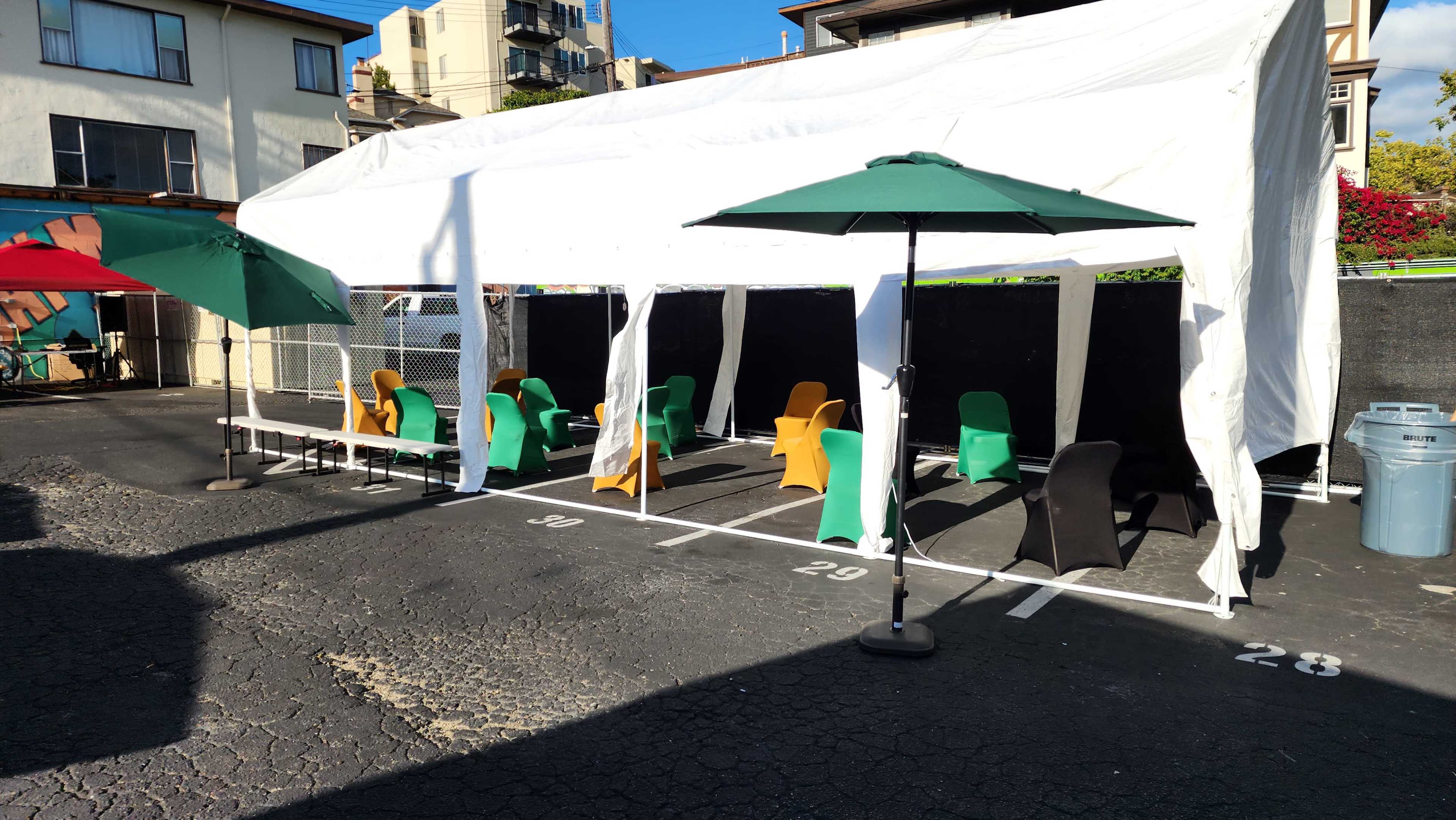 A white tent with green and red umbrellas covers several chairs arranged in a parking lot.