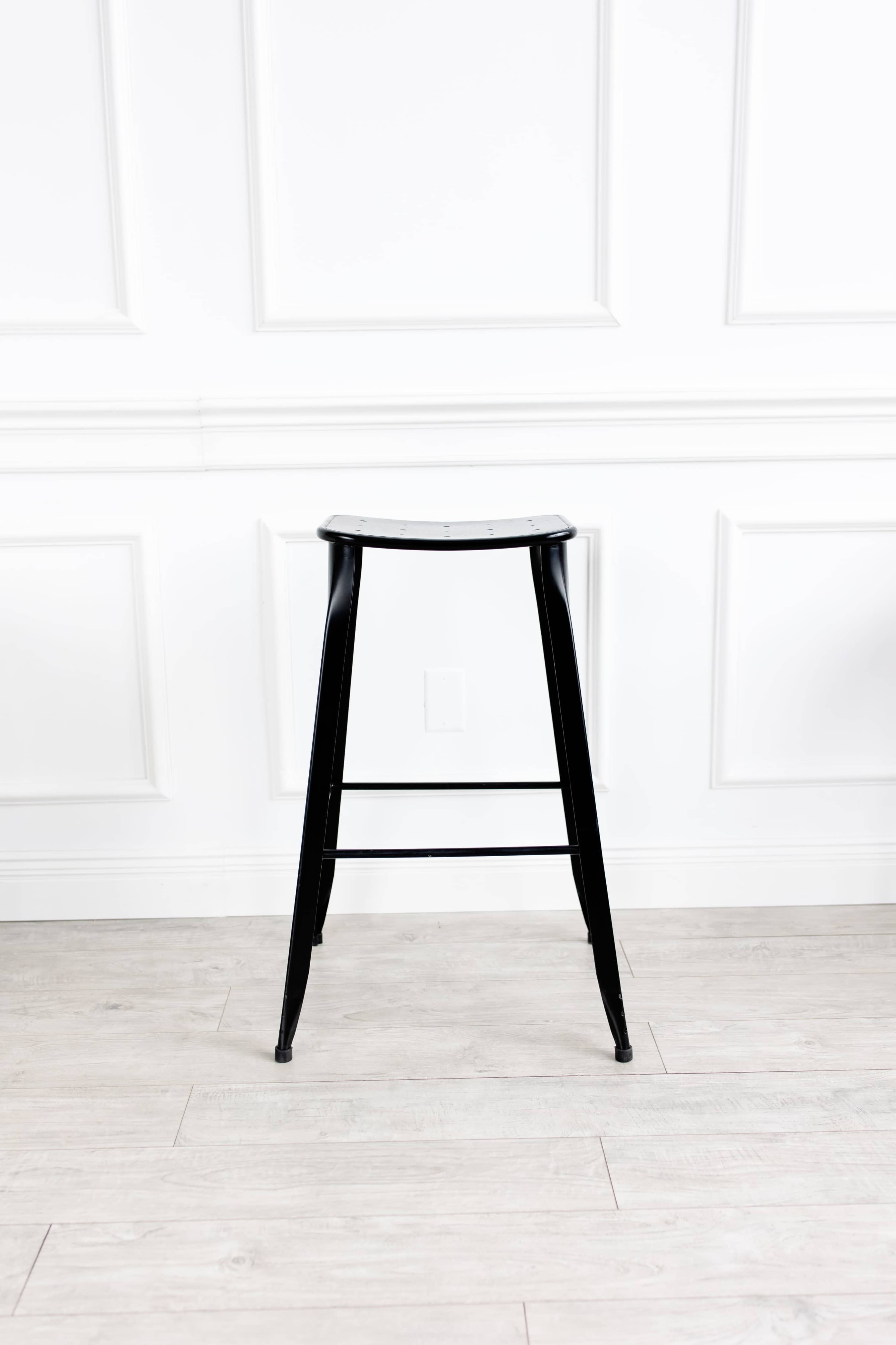 A black metal barstool stands alone against a white paneled wall with light-colored flooring.