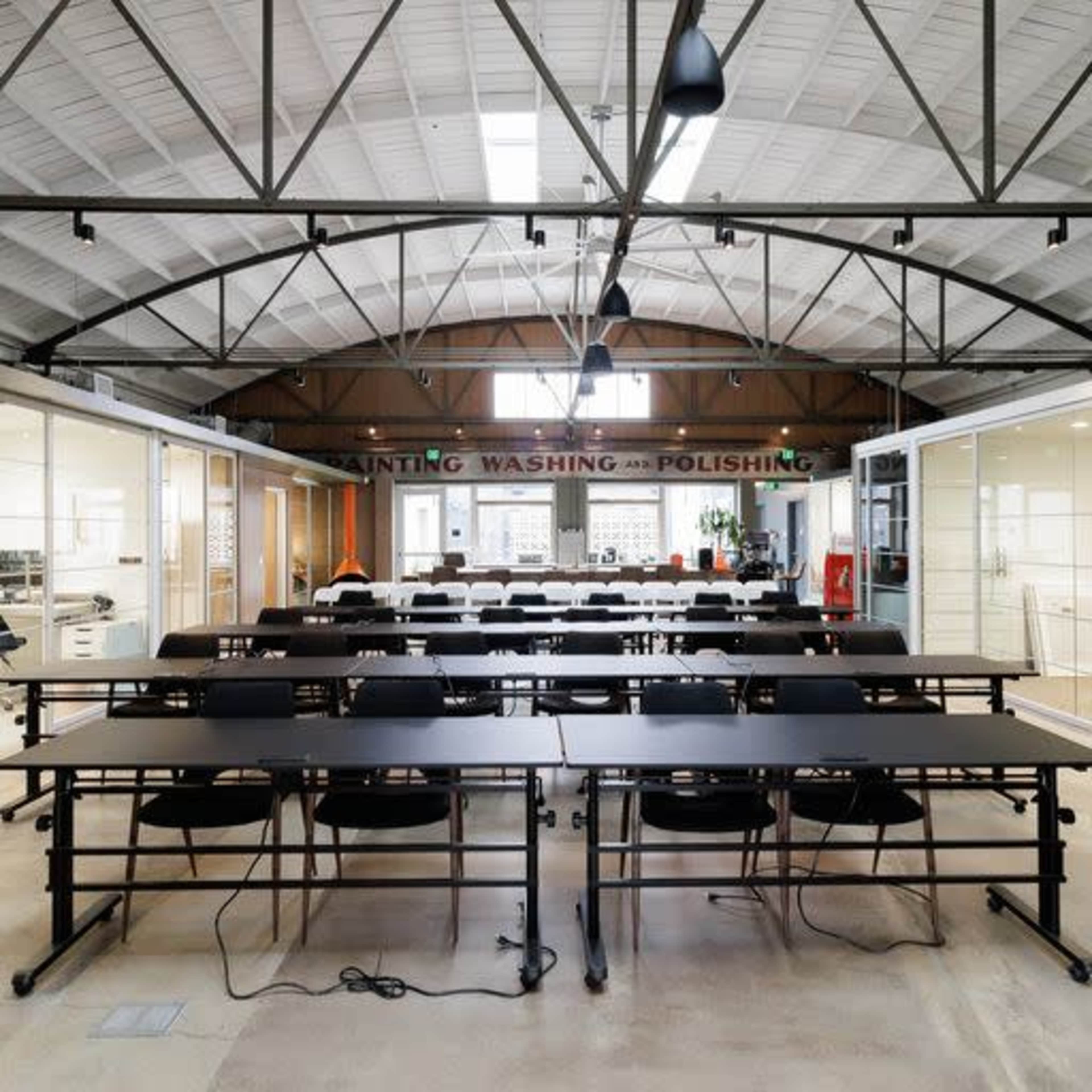 A spacious room features rows of long tables and chairs under a high vaulted ceiling with large windows.