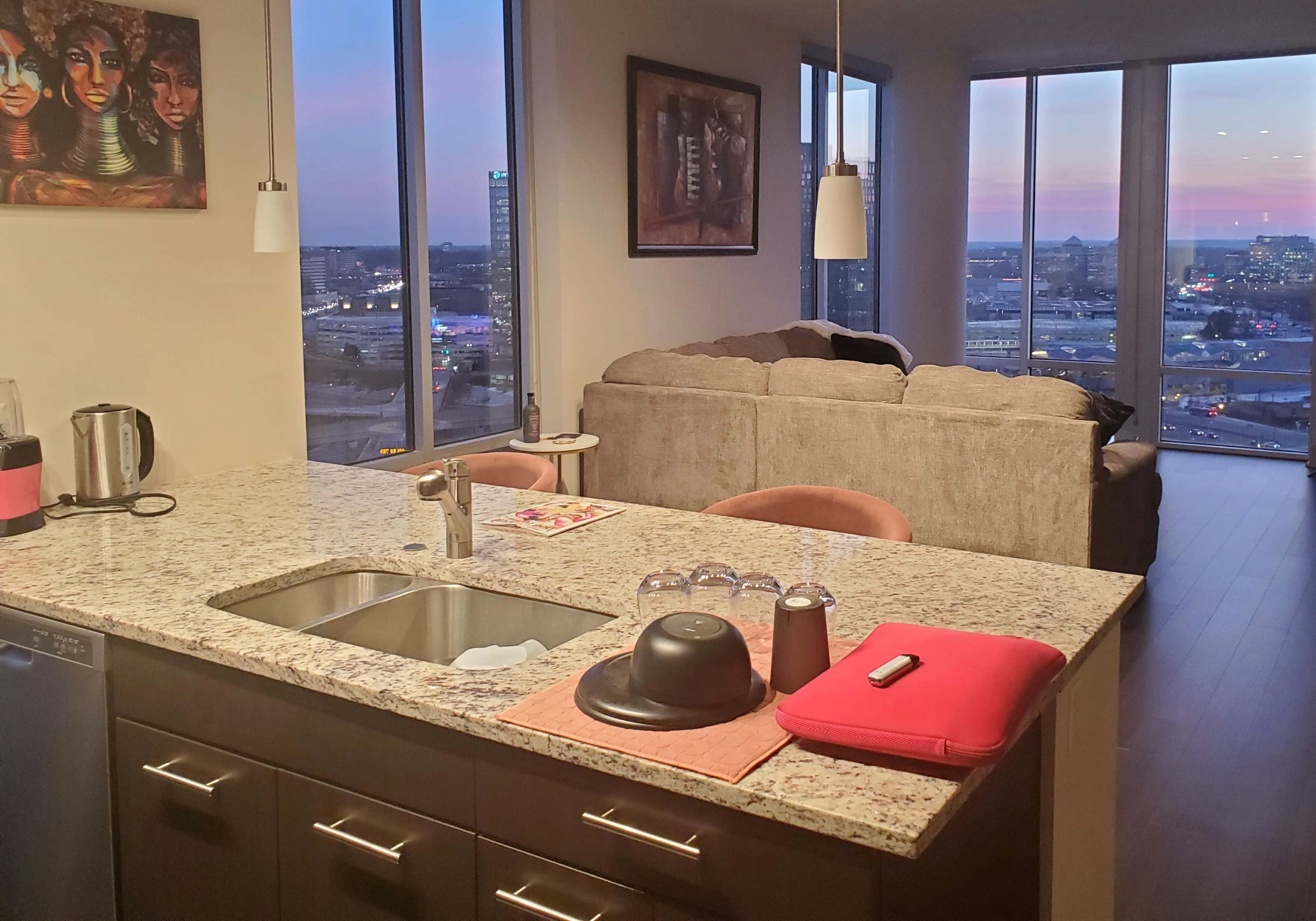 A modern kitchen with a granite countertop features a stainless steel sink and a view of a city skyline through large windows in a cozy living room setting.