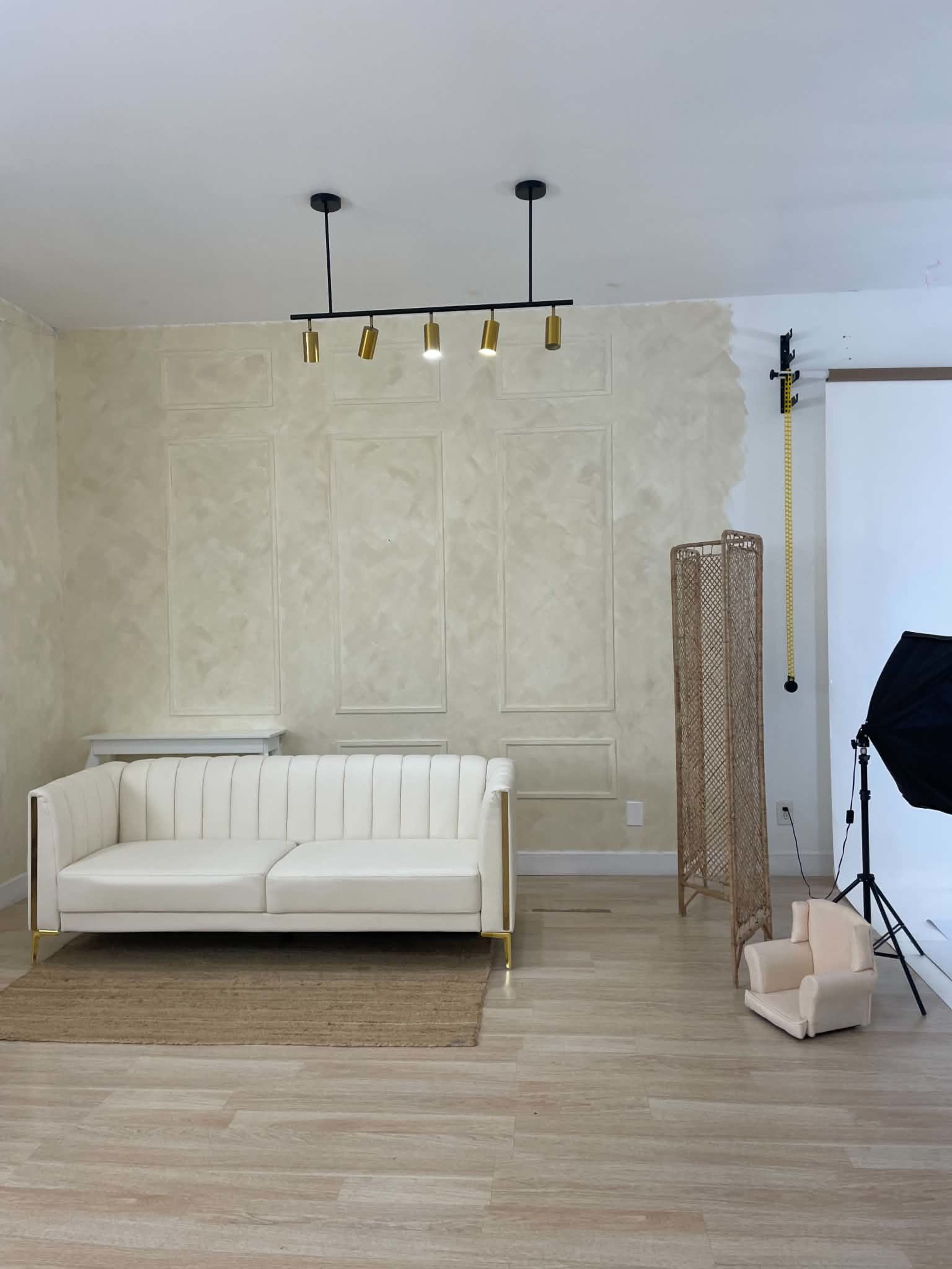The image shows a minimalist studio space featuring a white sofa, a decorative screen, a lighting setup, and a photography backdrop.