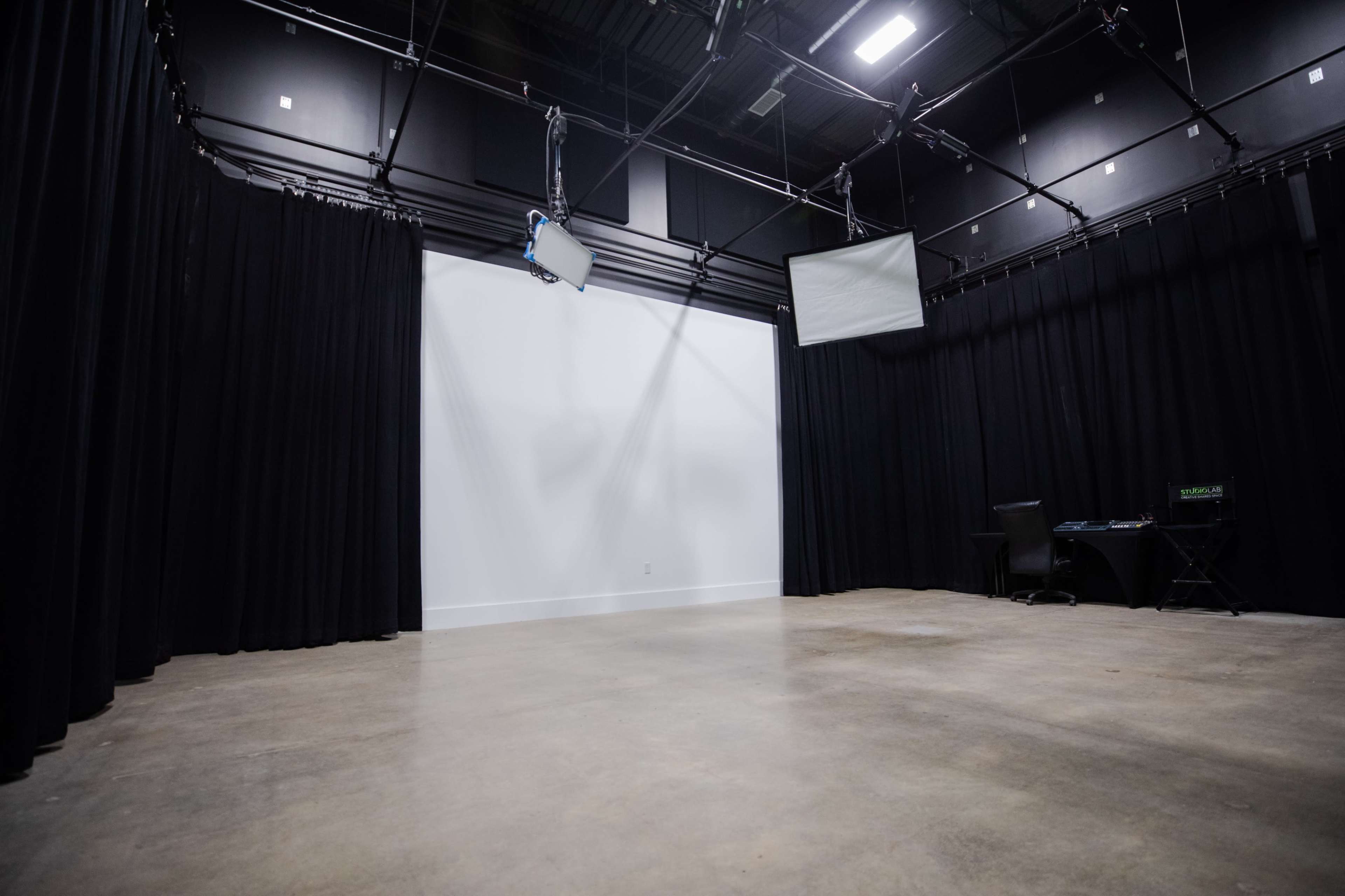 A bare studio space with black curtains, a large white wall, and two screens mounted on the ceiling.