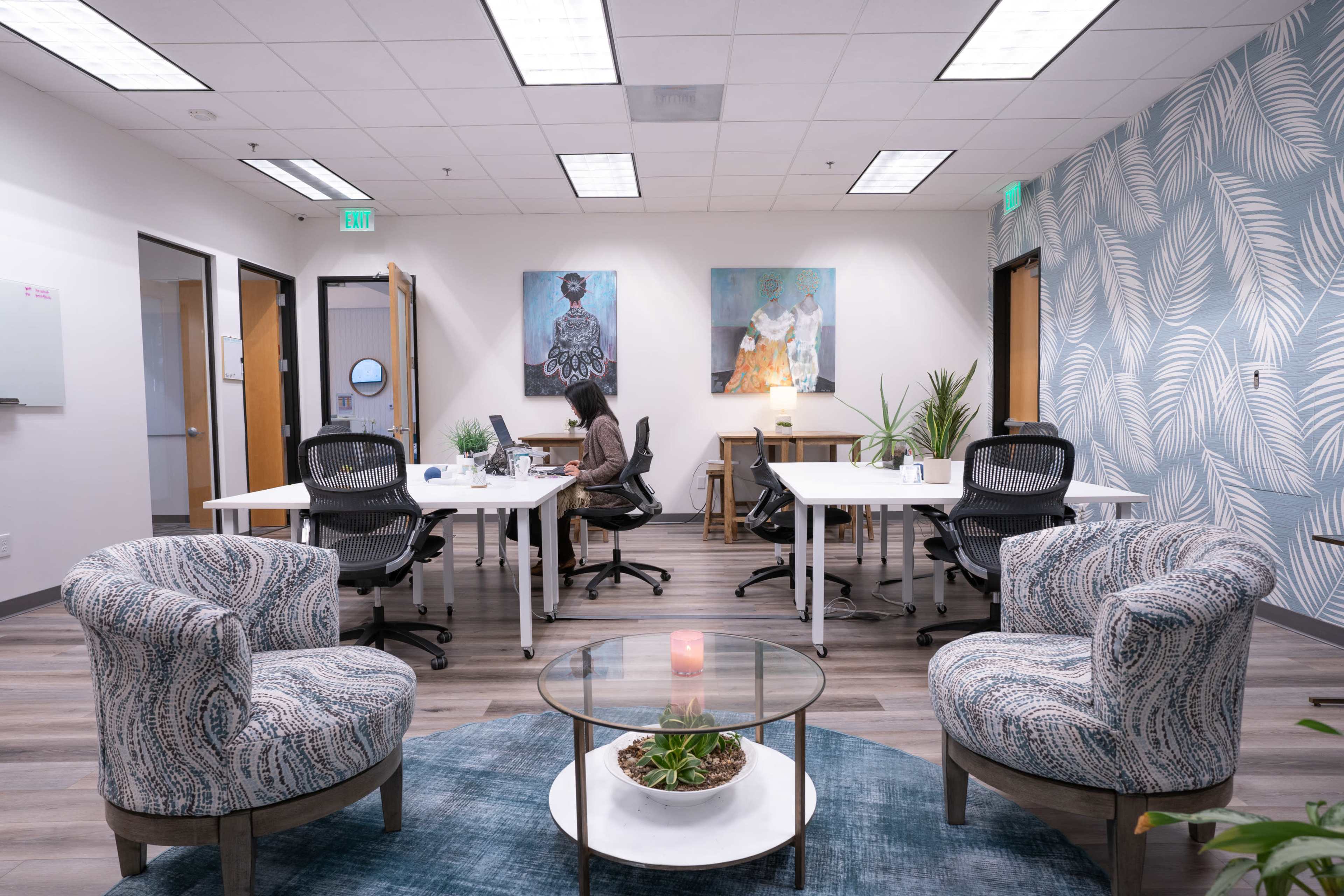 A modern office space features two seating areas with patterned chairs, a round table, and individual workstations equipped with computers against a backdrop of artwork and decorative plants.