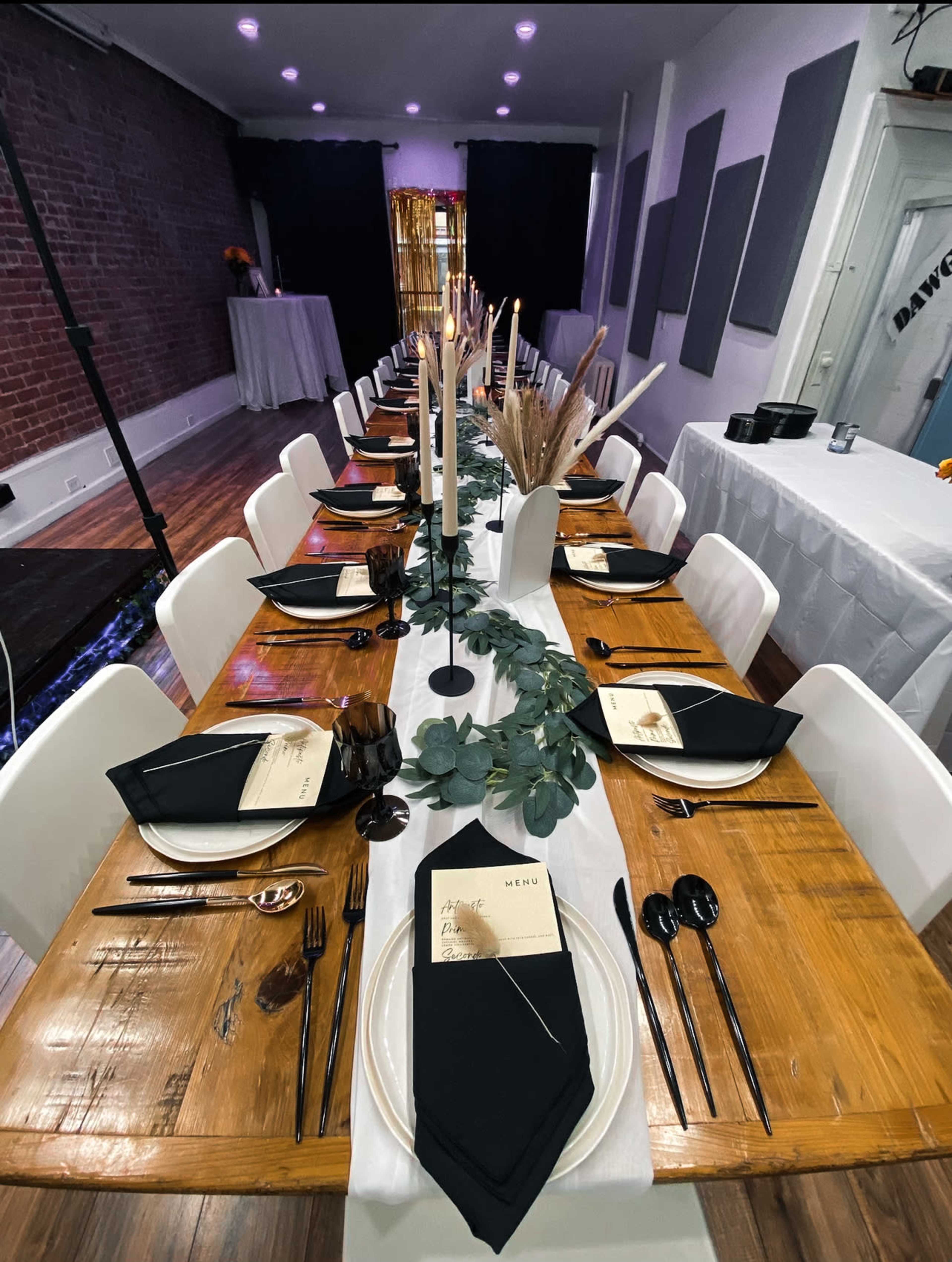 A long dining table is elegantly set with black napkins, plates, and utensils, featuring candles and greenery as centerpieces in a well-lit room.