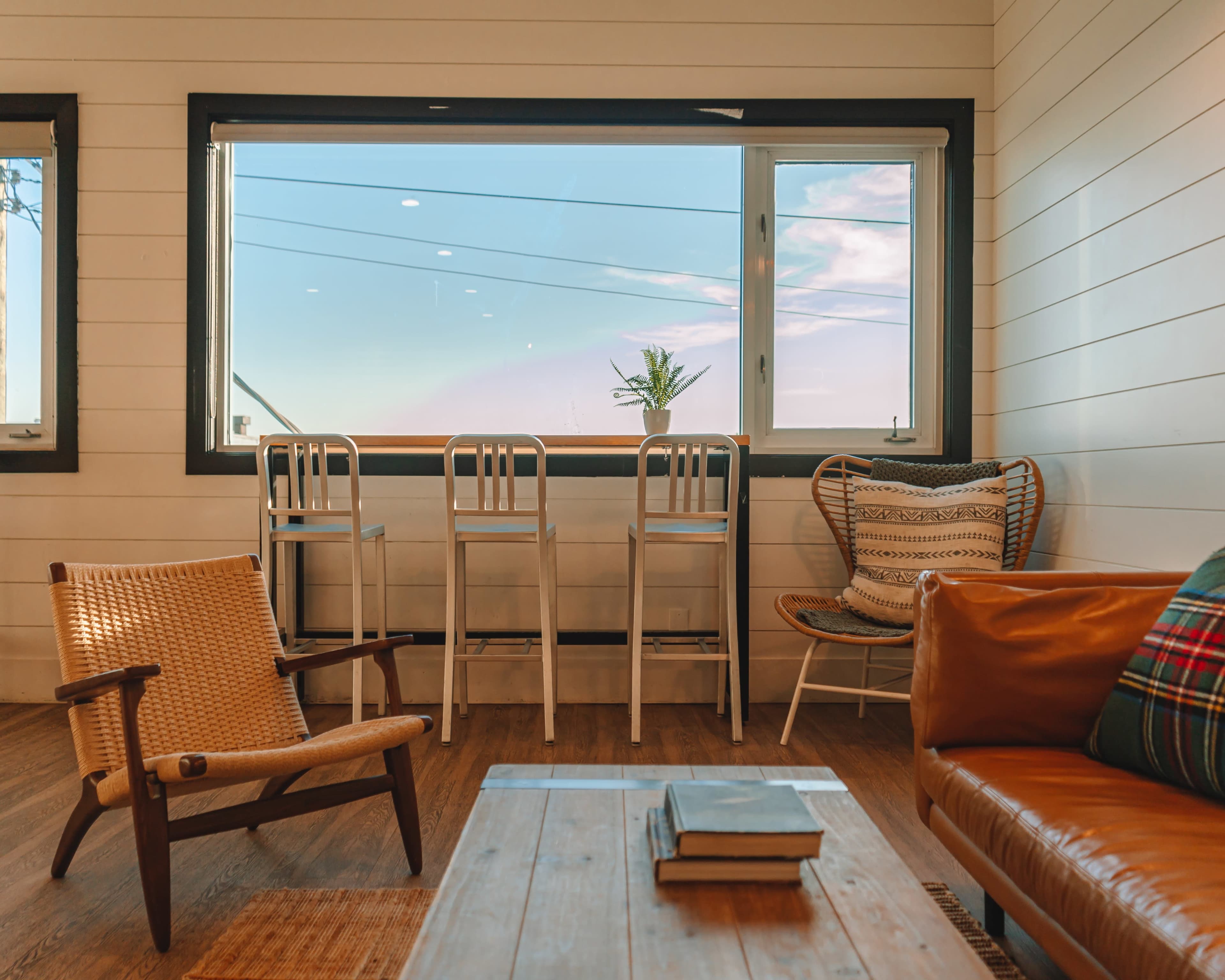 A cozy interior space featuring a wooden coffee table, a leather couch, a woven chair, and tall bar stools positioned by a large window.