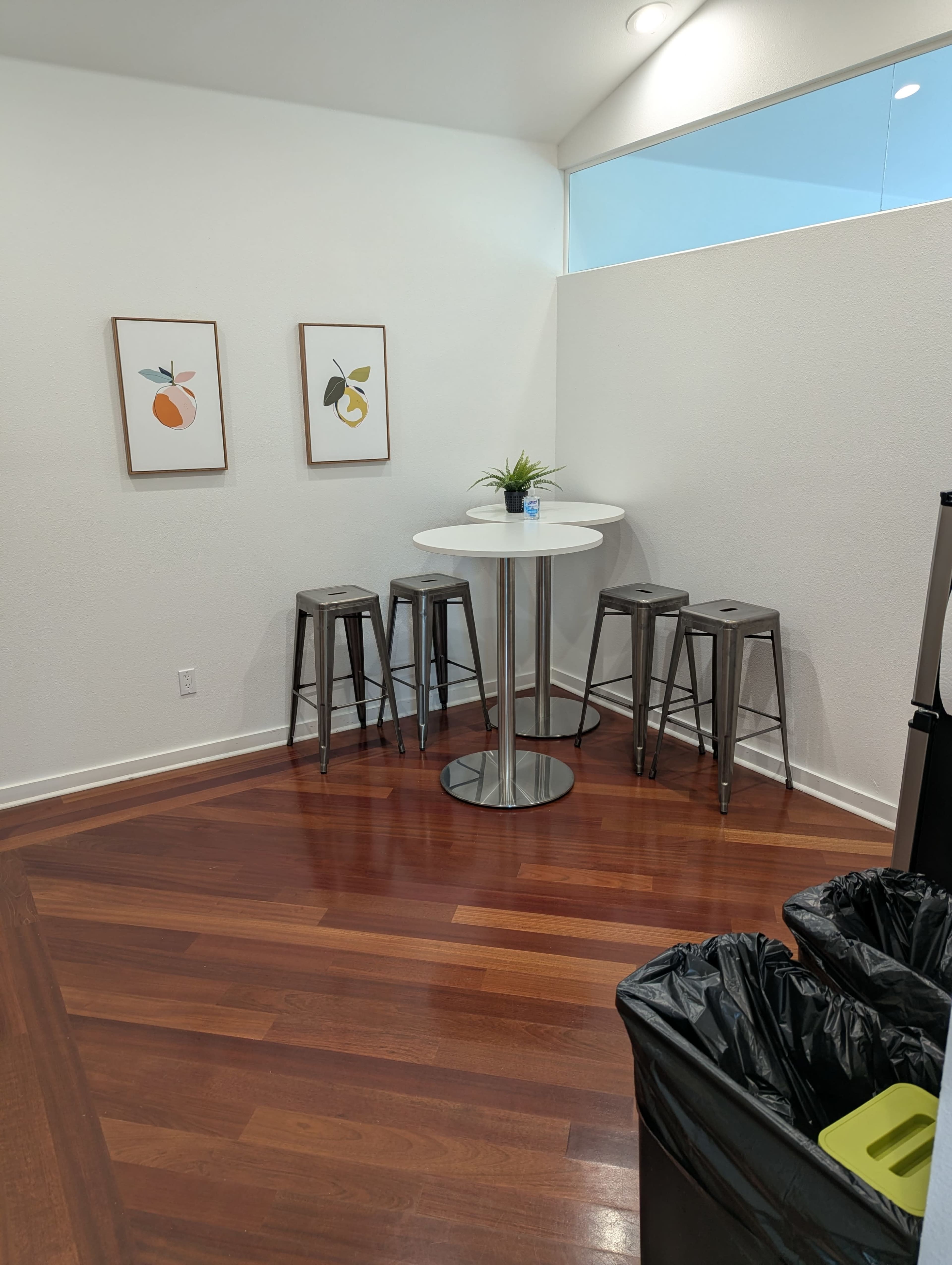 The image shows a small dining area with a round table and four metal stools, accompanied by two framed fruit prints on the wall and a trash bin in the corner.
