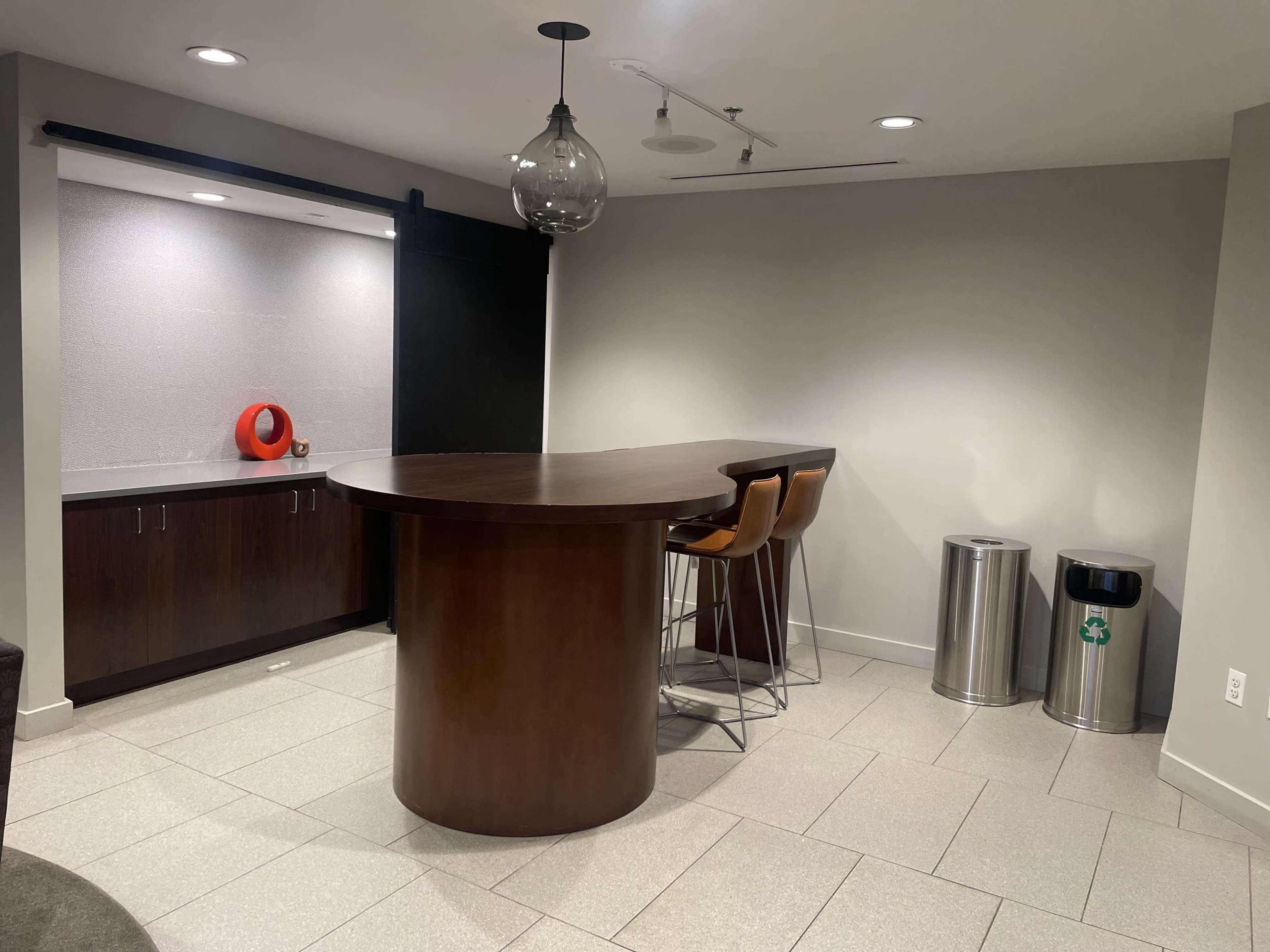 A modern kitchen space with a curved wooden bar, three high chairs, a minimalist countertop, and two trash bins.