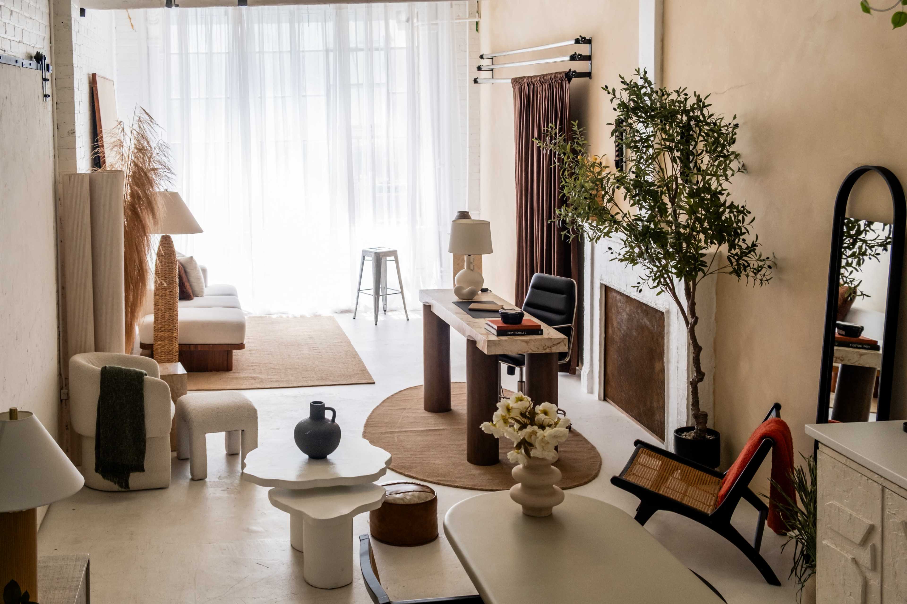 A spacious, well-lit interior features a mix of modern furniture, decorative plants, and soft textiles against a neutral backdrop.