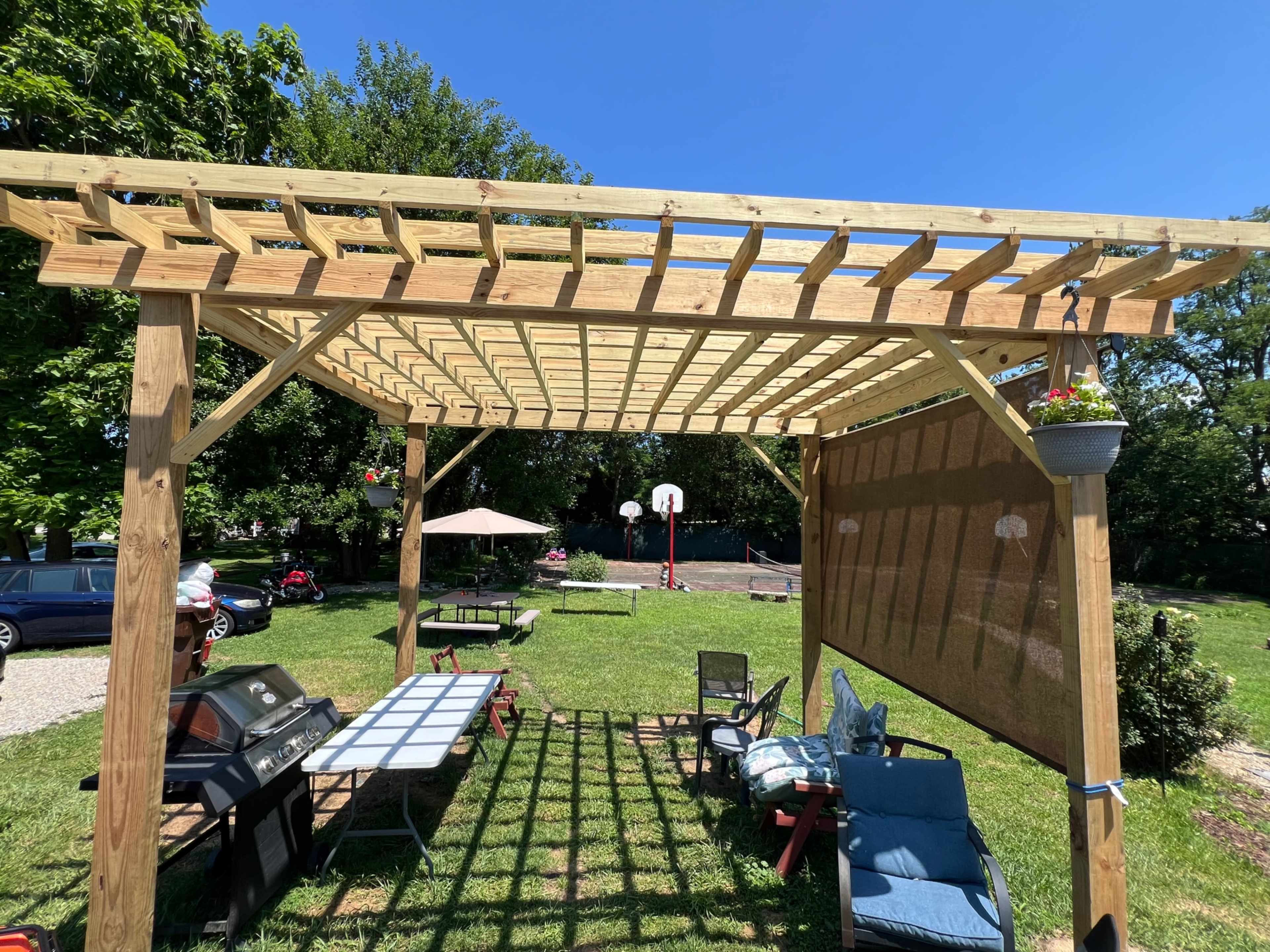 A wooden pergola stands in a grassy area, with a grill, tables, and chairs underneath, and a basketball court visible in the background.