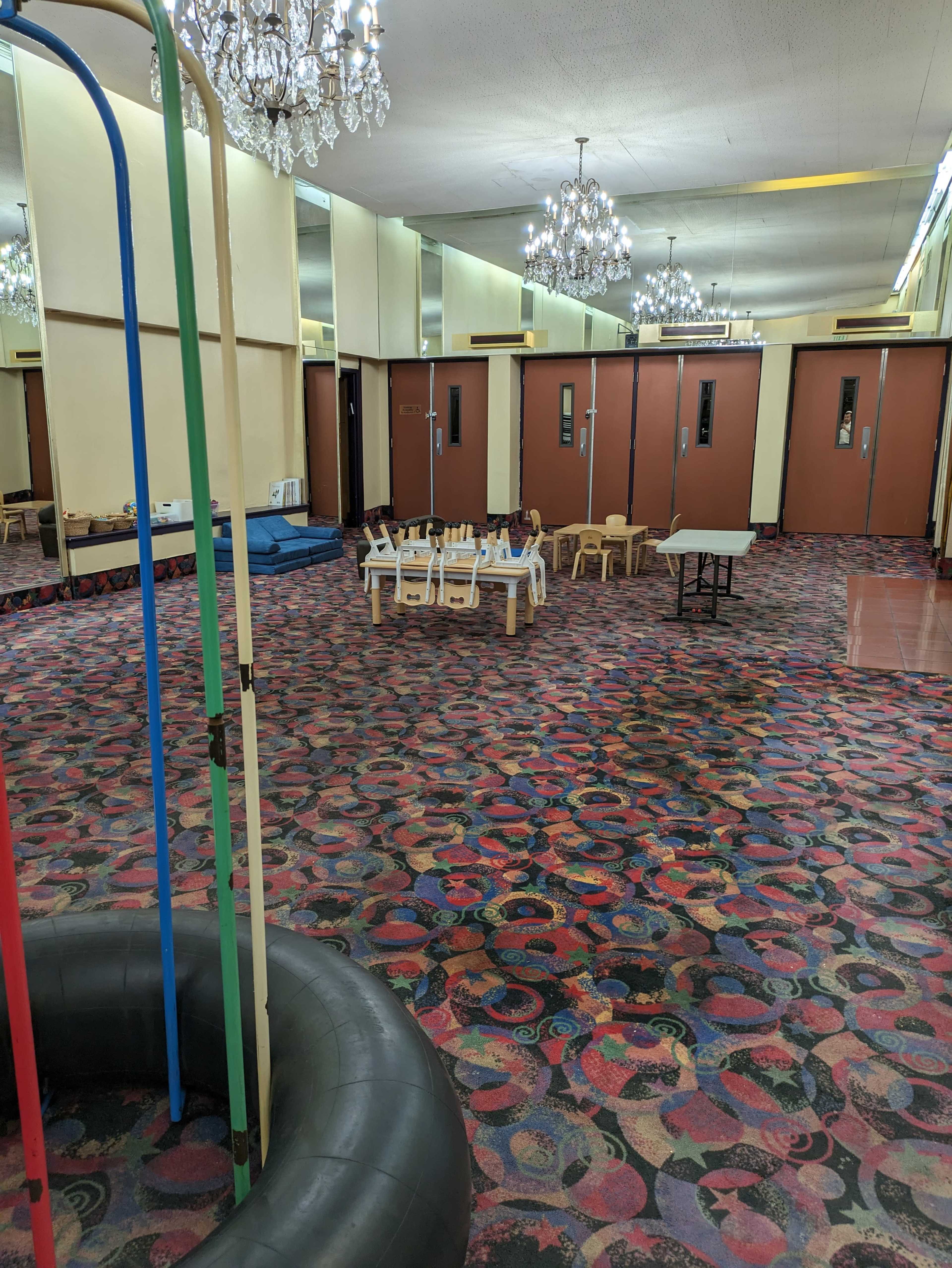 A spacious indoor area with colorful patterned carpeting, several small tables and chairs arranged in the center, and large mirrors lining the walls.