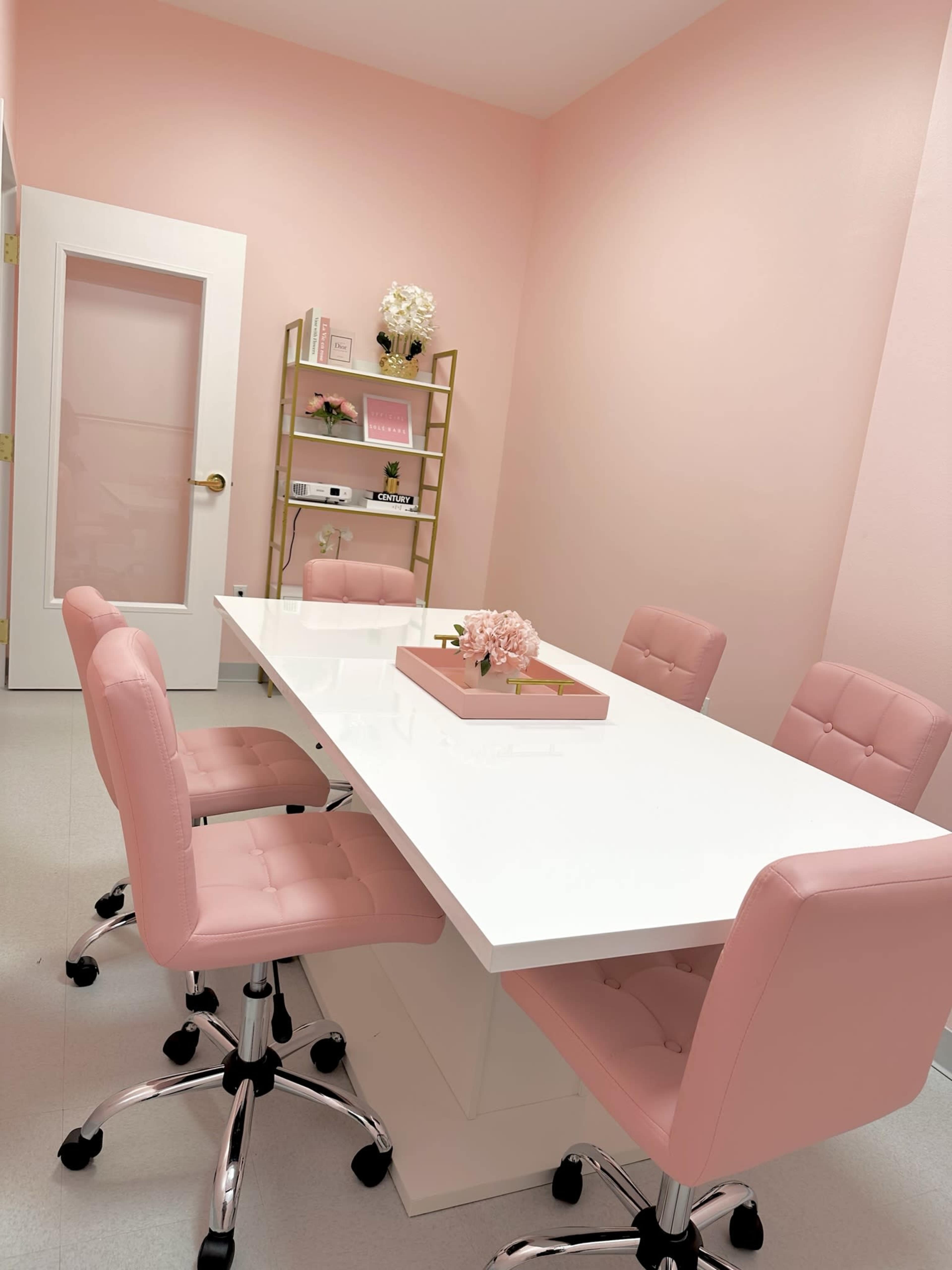 A pink-themed meeting room with a white table surrounded by four pink chairs and a shelving unit against the wall.