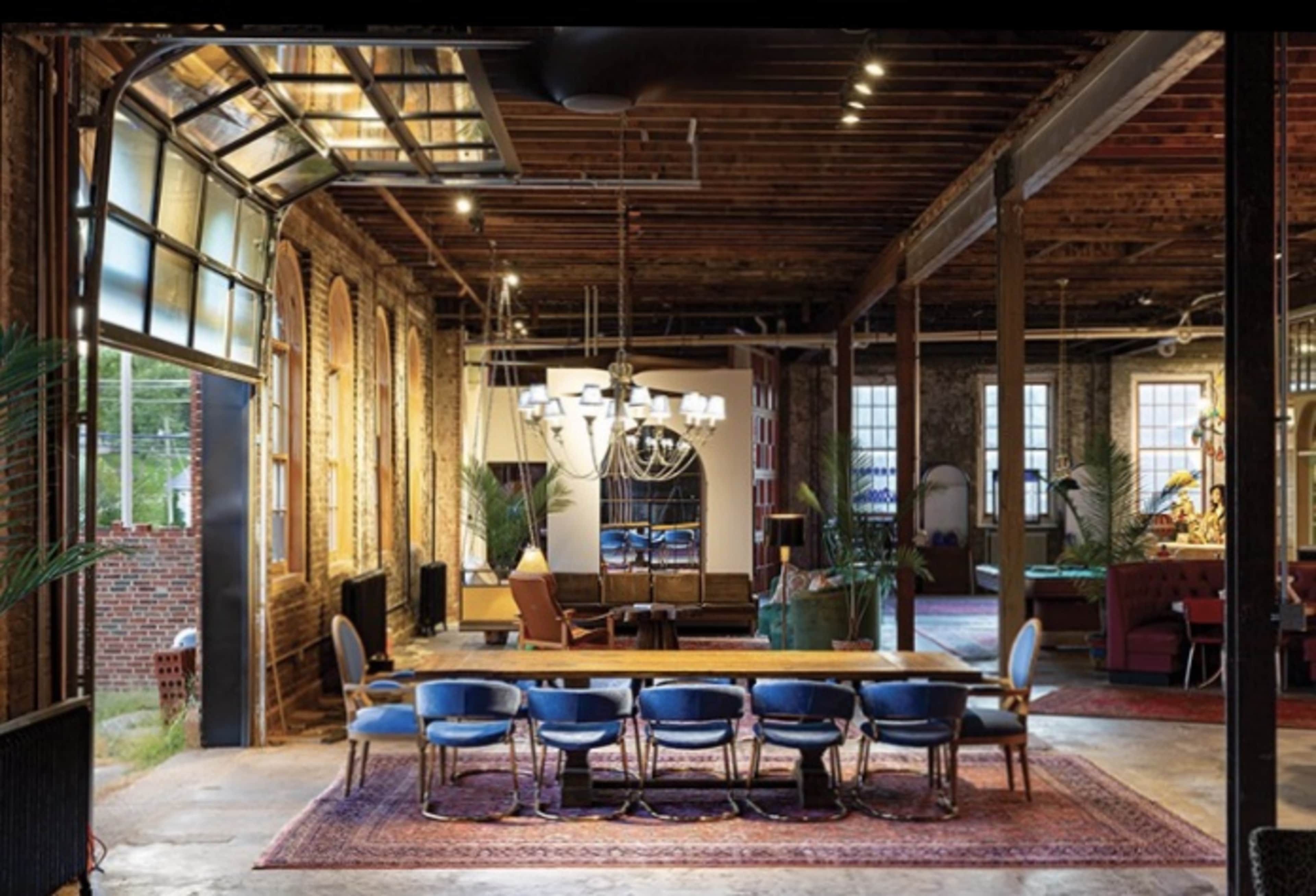 The image shows a spacious, industrial-style interior featuring a long table surrounded by chairs, large windows, and exposed wooden beams in the ceiling.